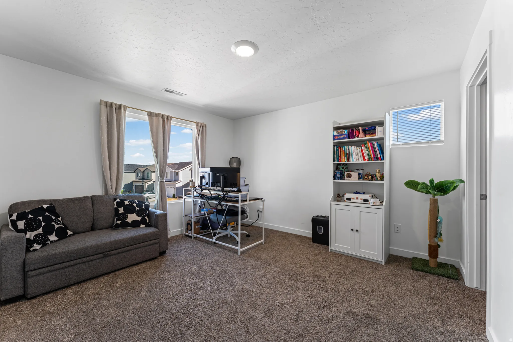 Carpeted room featuring a large window with light-filtering drapes, a built-in bookshelf with lower cabinetry, and a ceiling-mounted light fixture