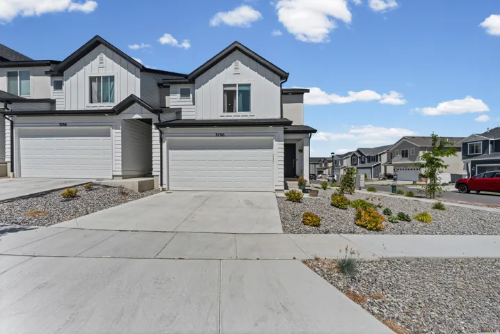 Modern townhome exterior featuring vertical white siding, dark trim, an attached garage, paved driveway, and low-maintenance landscaping with rock beds and shrubs