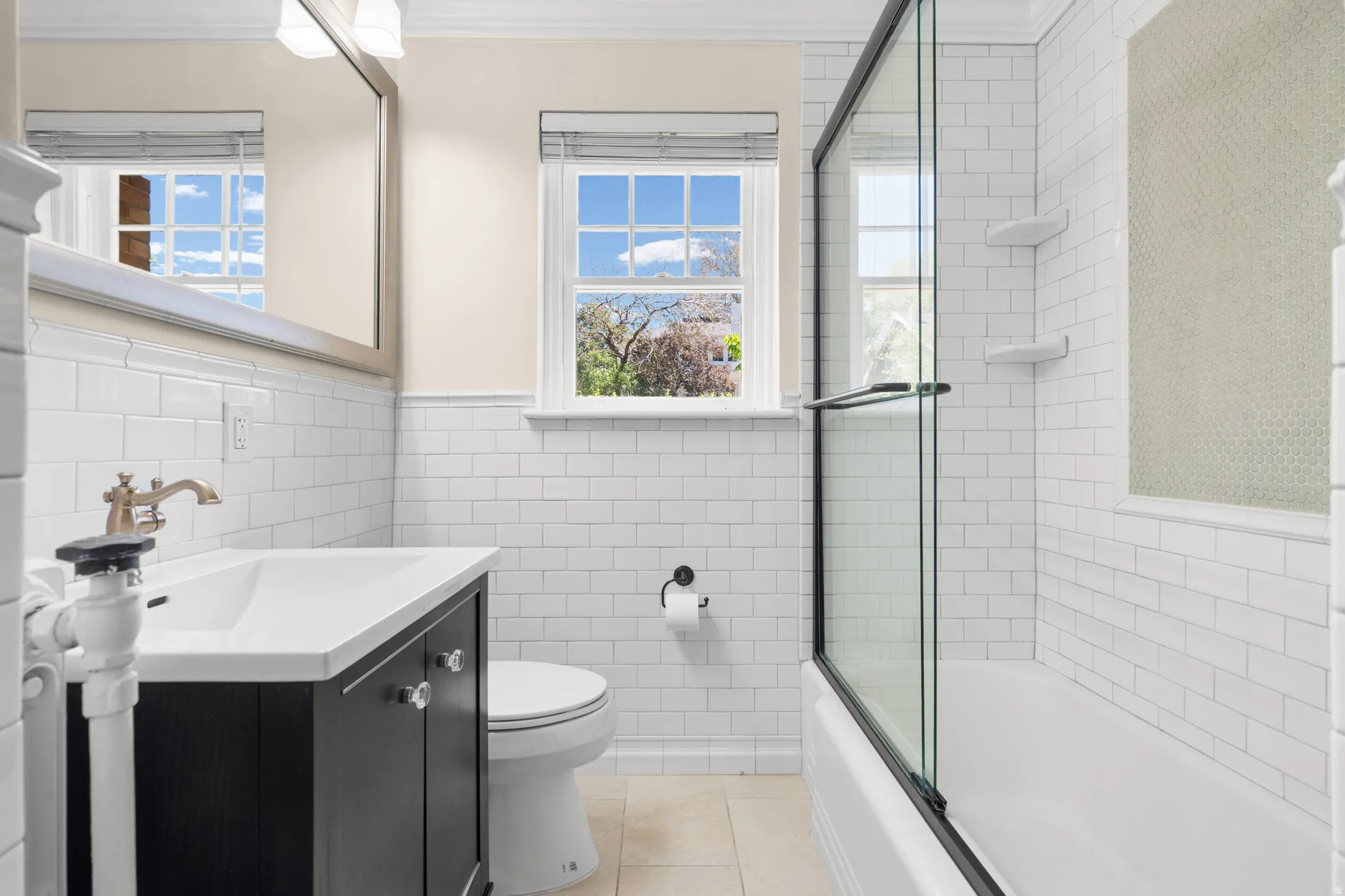 Bathroom featuring white subway tile walls, a vanity with a white countertop and brass-finish faucet, an integrated bathtub and shower with a glass enclosure, and a window for natural light