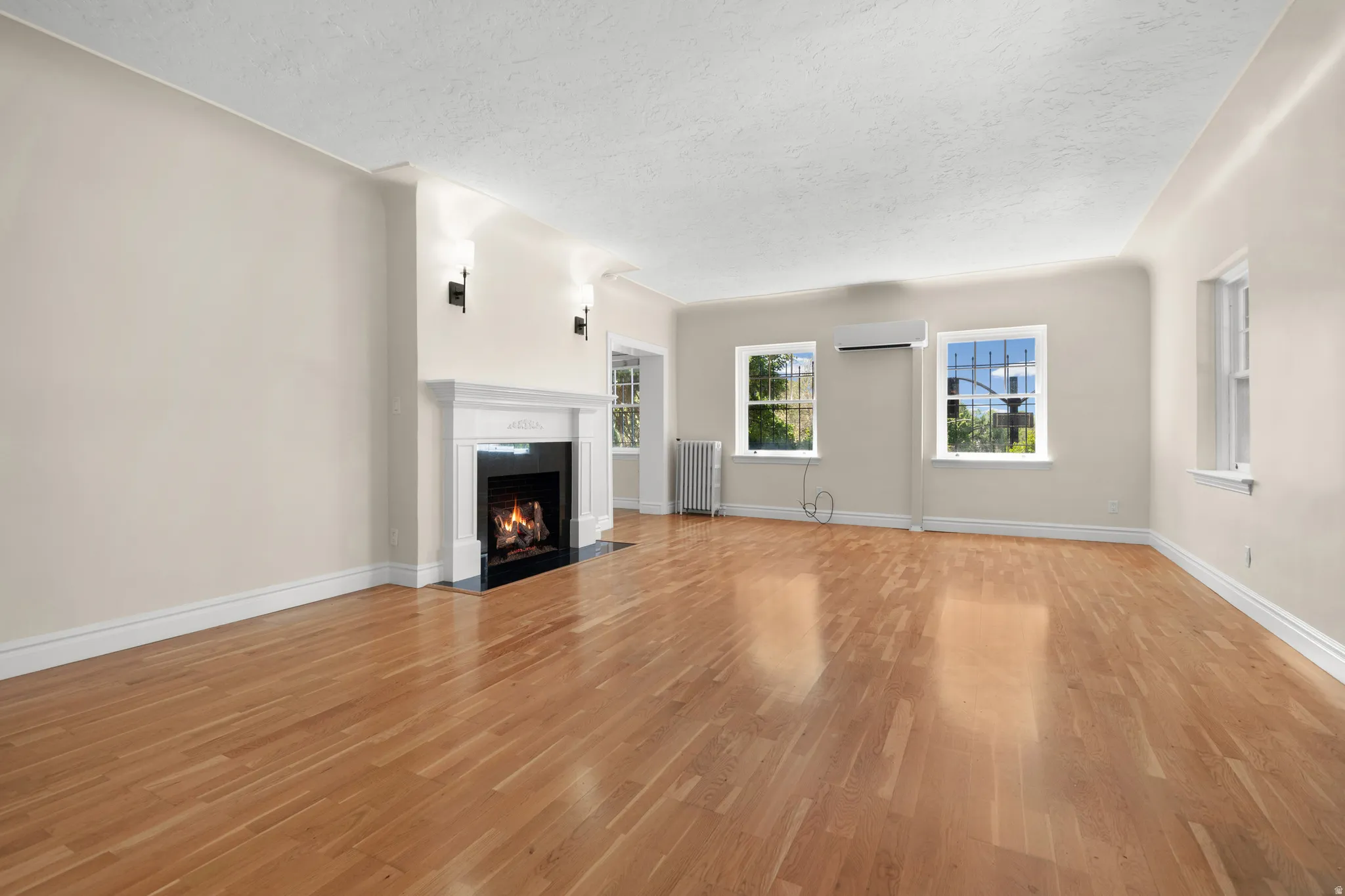 Spacious room featuring wood-finish flooring, a fireplace with a white mantel and black surround, three windows allowing ample natural light, and a wall-mounted air conditioning unit