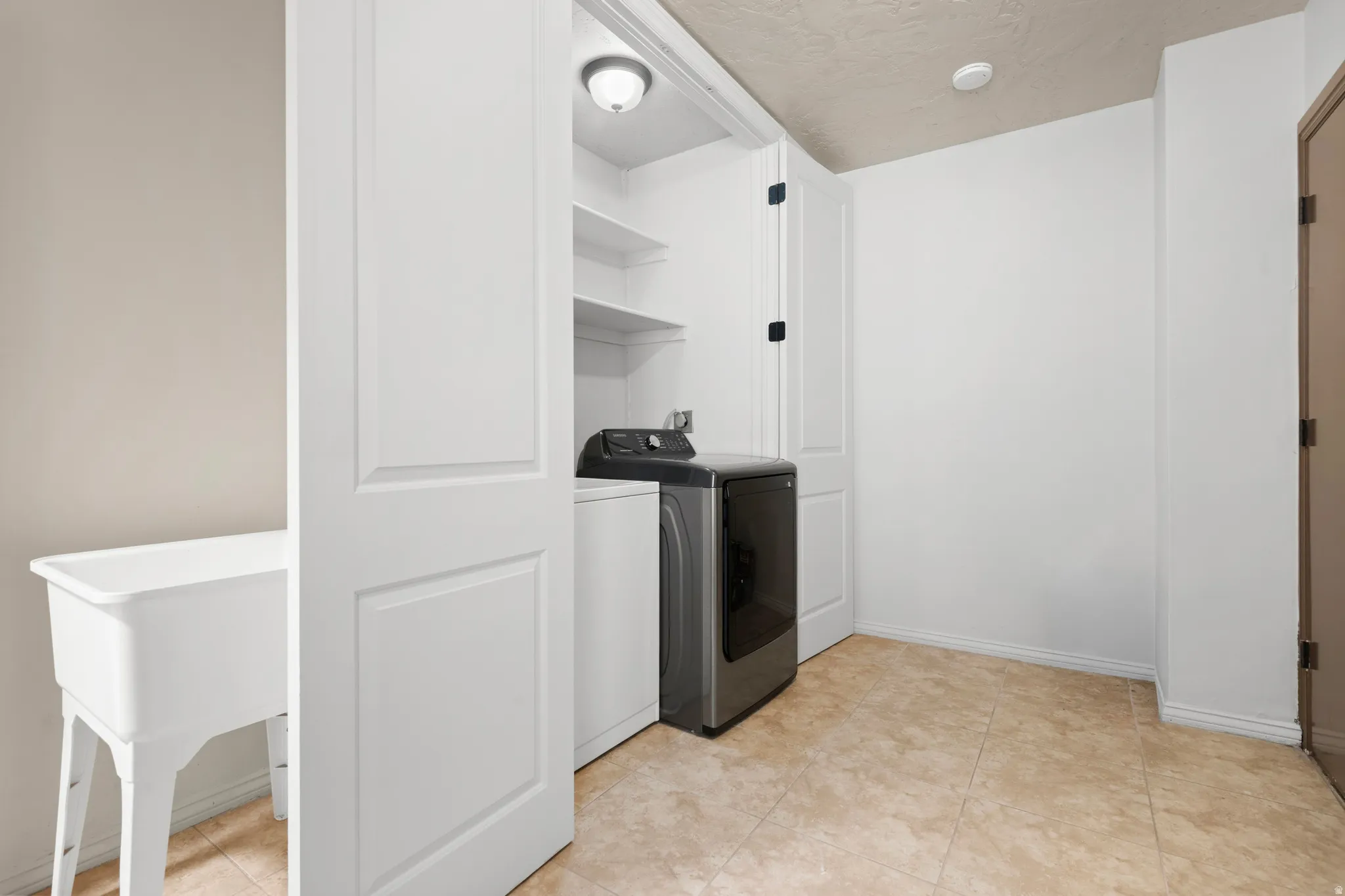Dedicated laundry area featuring a utility sink, light-colored tile flooring, overhead built-in shelving, and white paneled bi-fold doors