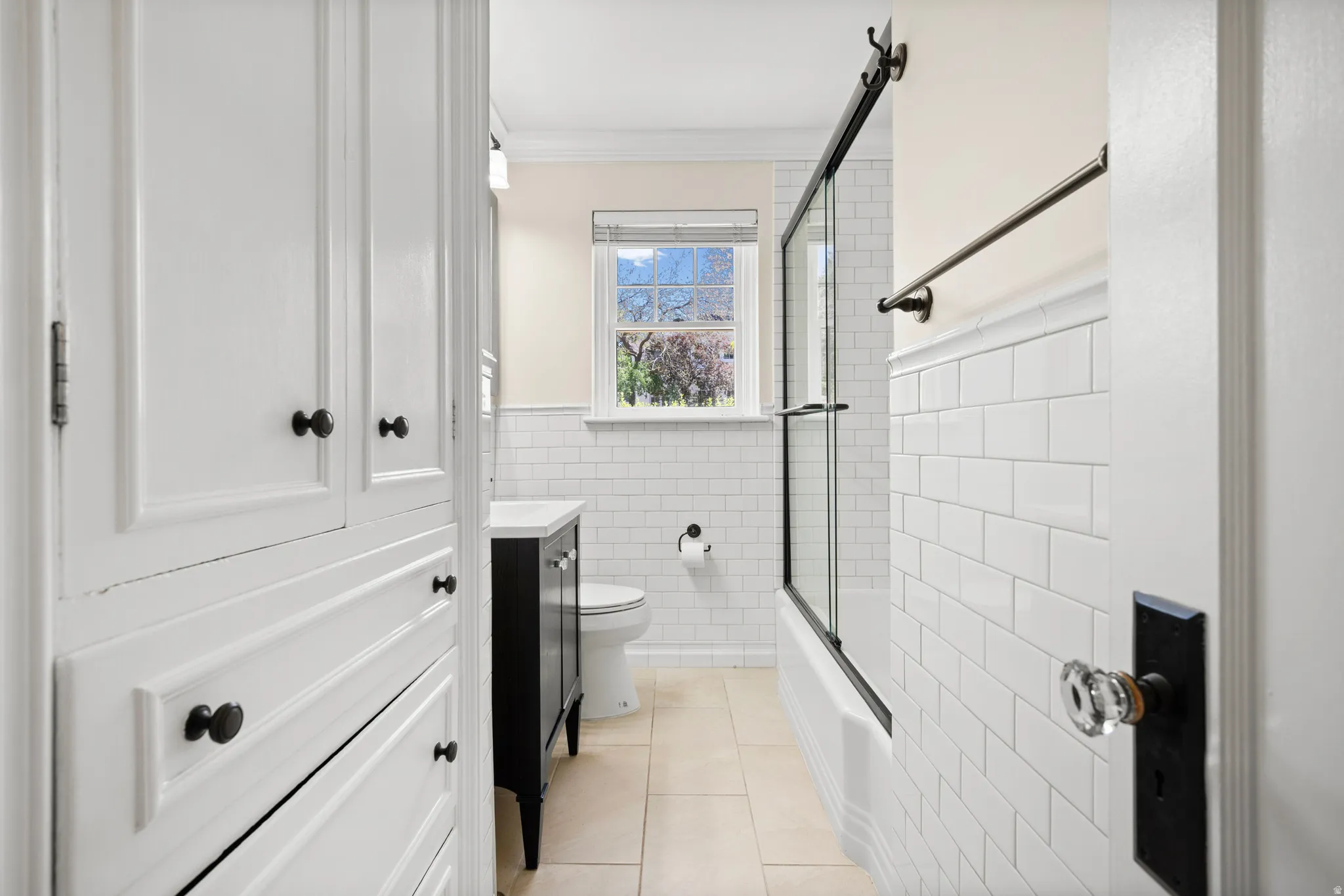 Bathroom featuring built-in cabinetry with black hardware, subway tile wainscoting, a black vanity with a white countertop, and a shower-over-tub with a framed glass enclosure