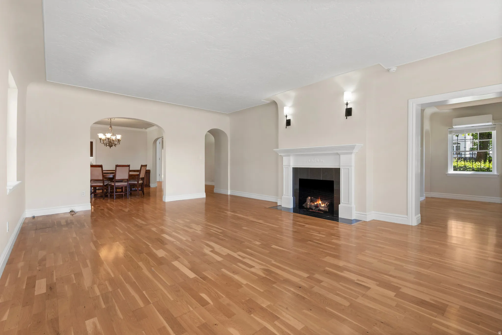Spacious living area featuring light wood-finish flooring, a classic white mantel fireplace with a black tile surround, and architectural archways