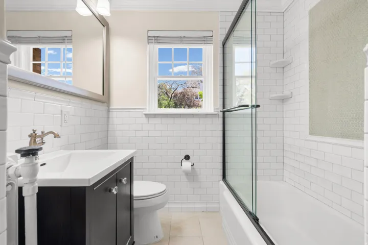 Bathroom featuring white subway tile walls, a vanity with a white countertop and brass-finish faucet, an integrated bathtub and shower with a glass enclosure, and a window for natural light