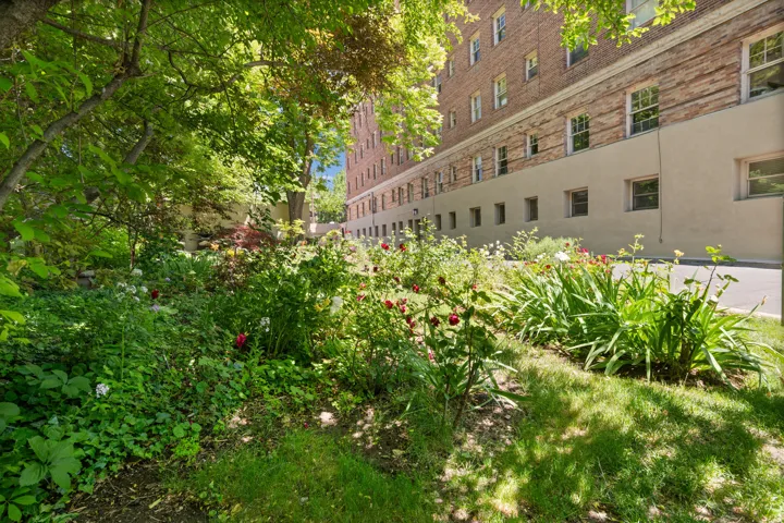 Lush garden landscaping with mature trees and varied plantings, featuring a brick and stucco exterior building in the background