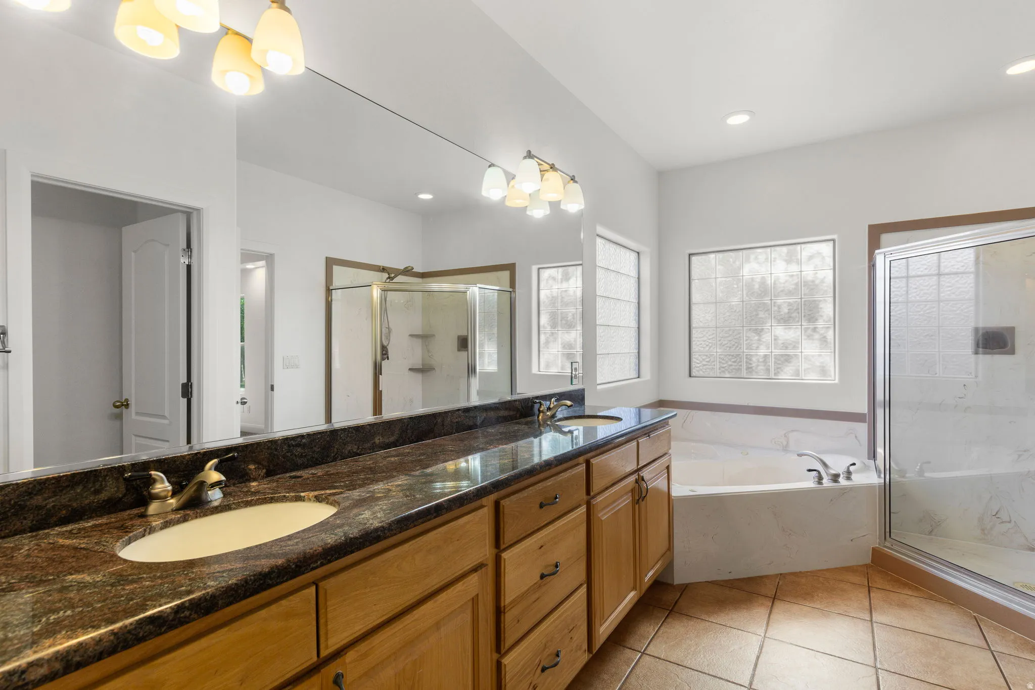 Spacious bathroom featuring a dual vanity with a patterned countertop, a large soaking tub, a separate glass-enclosed shower, and decorative privacy windows