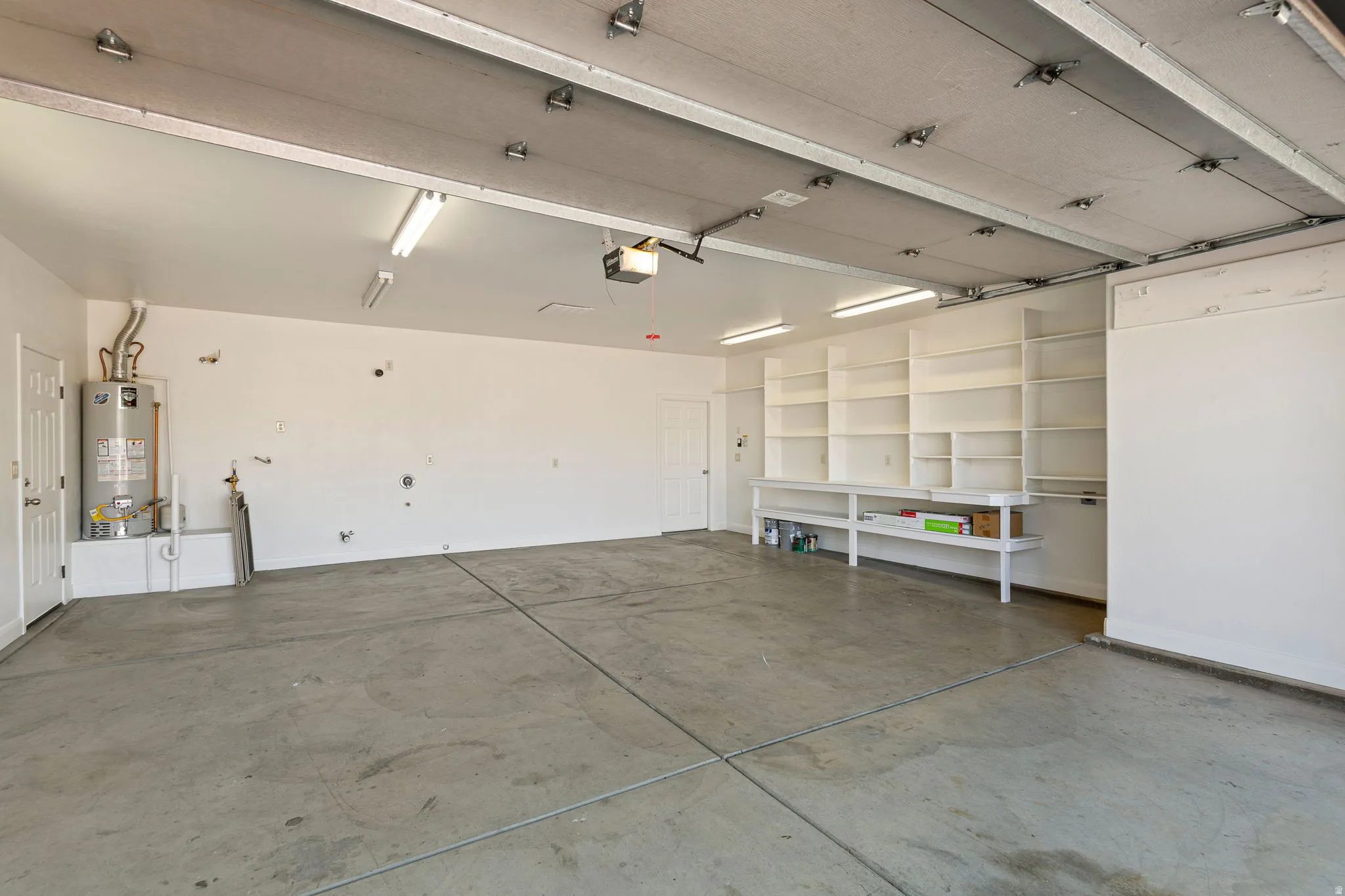 Spacious garage with concrete flooring, fluorescent lighting, built-in shelving, a workbench, and a water heater