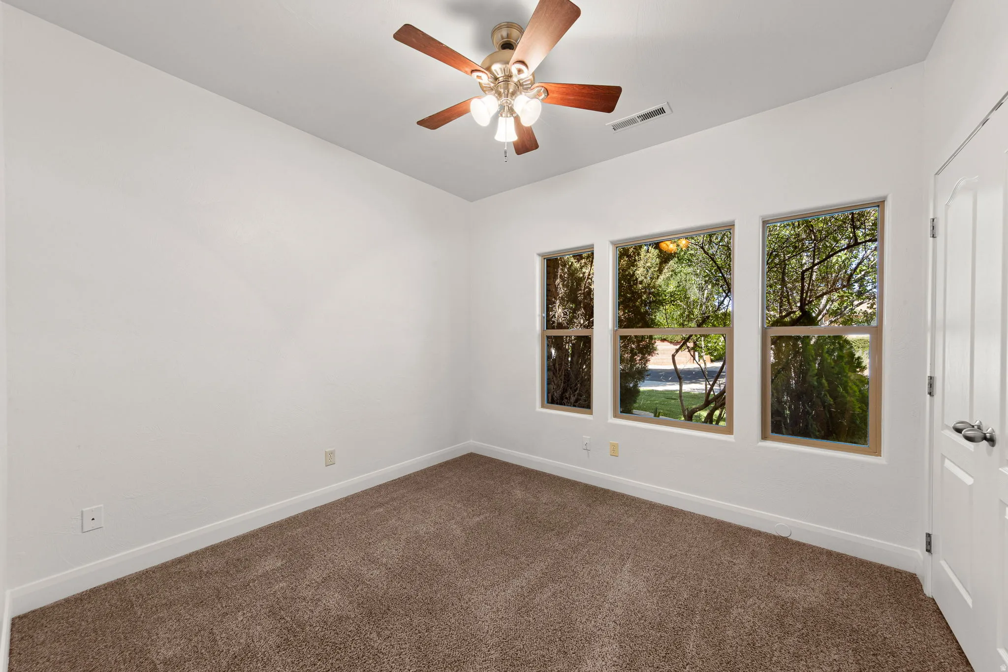 Room featuring three large windows, a ceiling fan with light fixture, carpeted flooring, and a paneled door with dark hardware