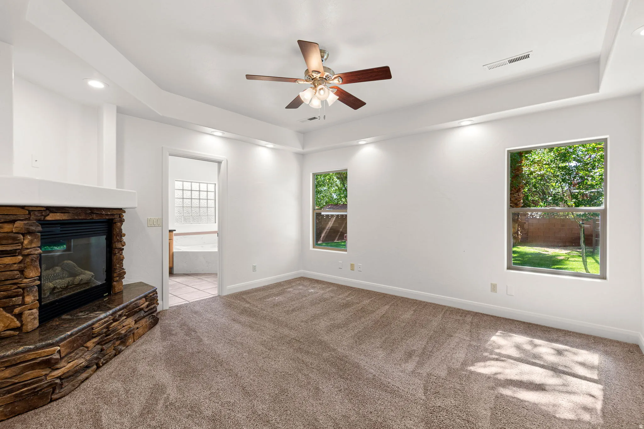 Spacious room featuring a stone-clad fireplace with a hearth, wall-to-wall carpeting, and a tray ceiling with recessed lighting