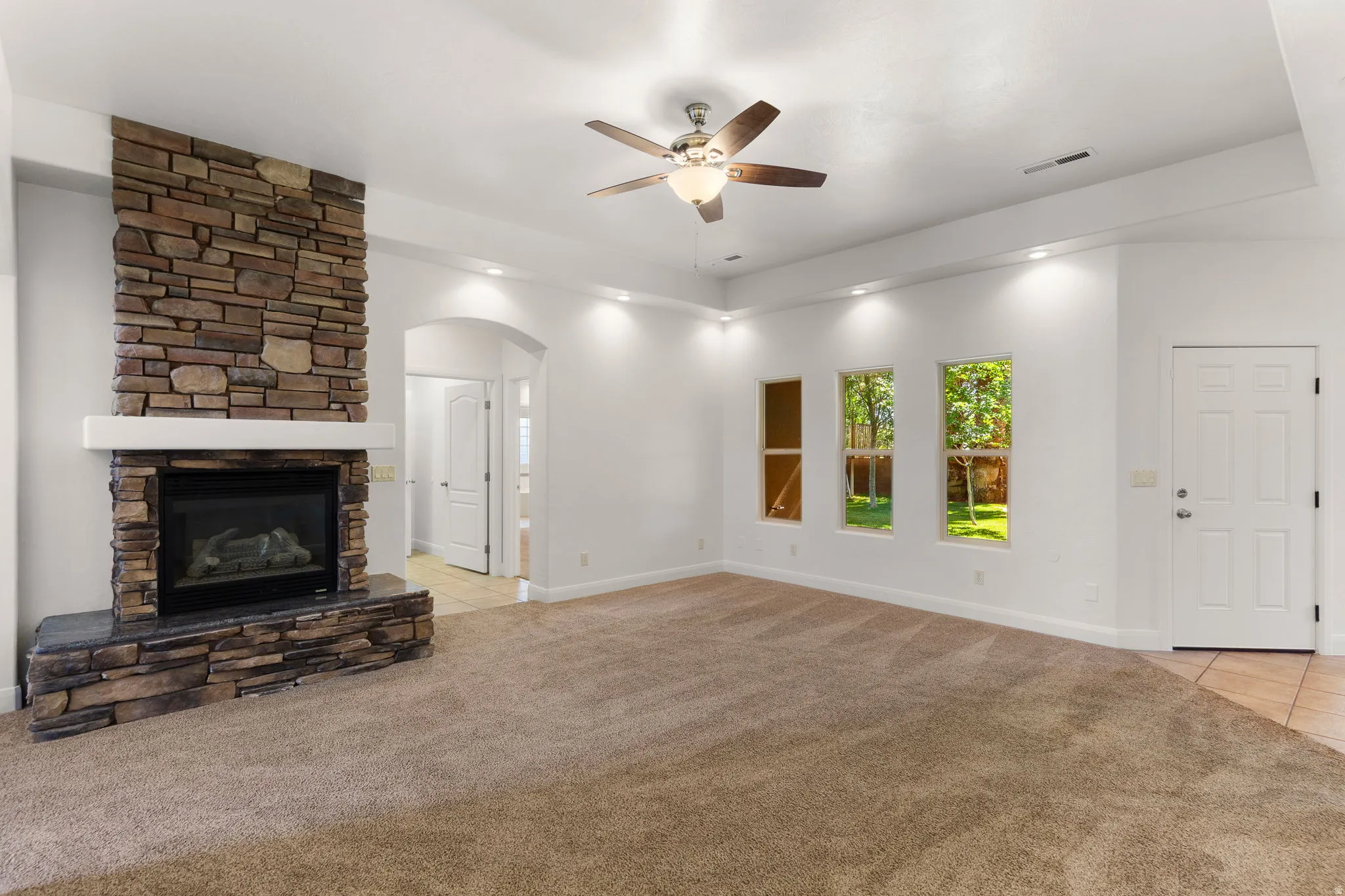 Spacious living area featuring a stone-clad fireplace with a hearth, a ceiling fan, recessed lighting, and ample natural light from multiple windows
