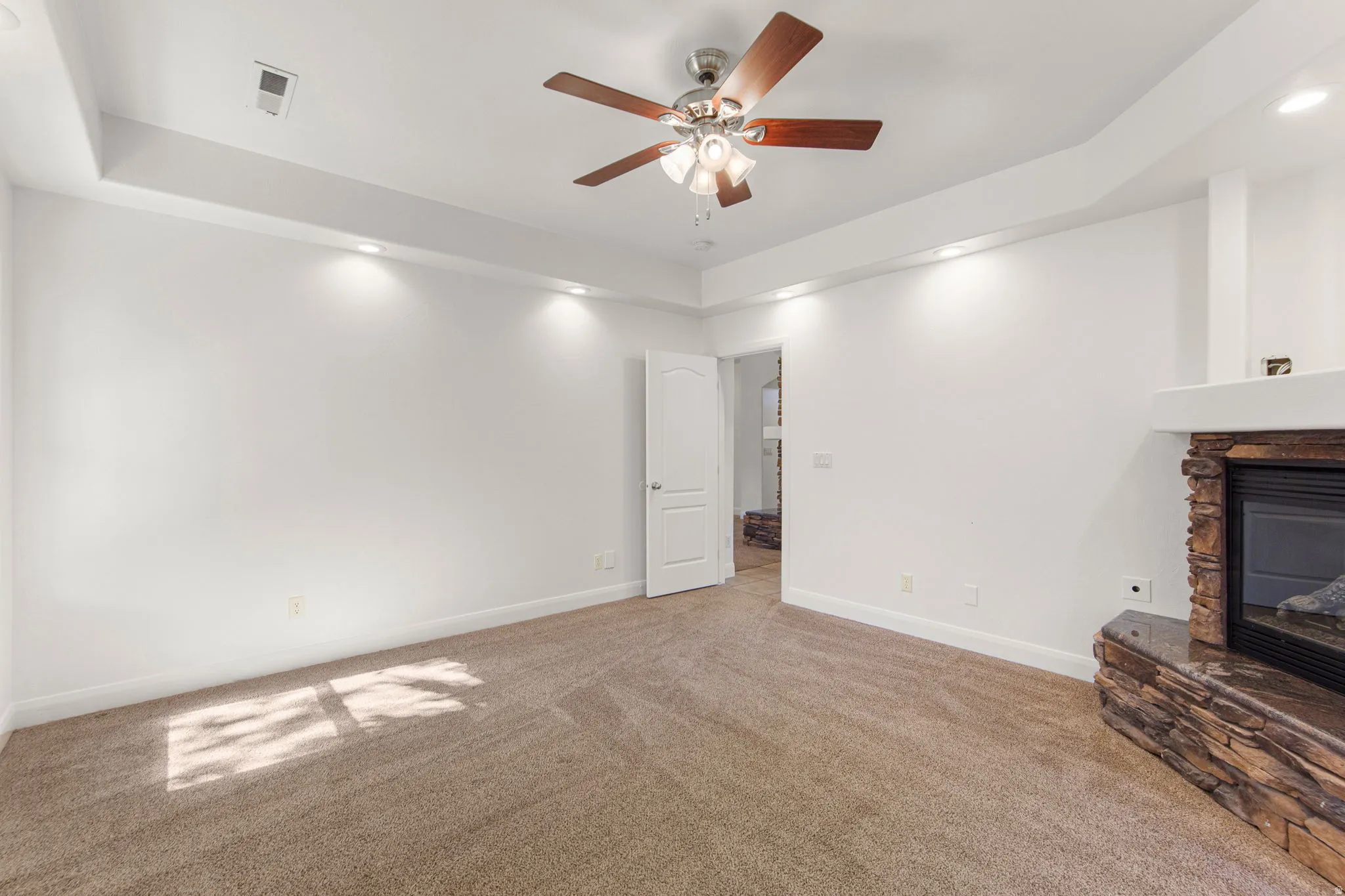 Carpeted interior space featuring a stacked stone fireplace with a dark stone hearth and mantle, a ceiling fan with integrated lighting, and recessed wall lighting