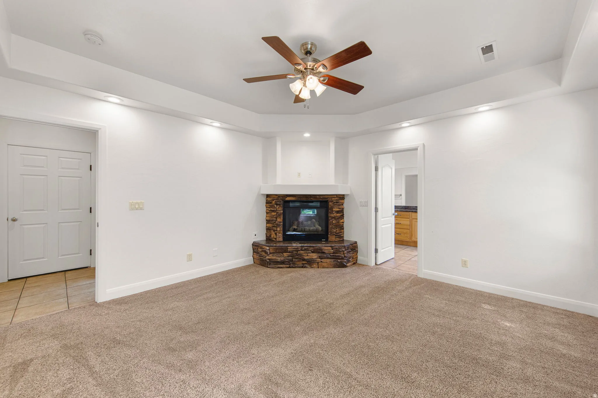 Spacious room featuring a stone-clad corner fireplace with a mantel, tray ceiling with recessed lighting, and a ceiling fan