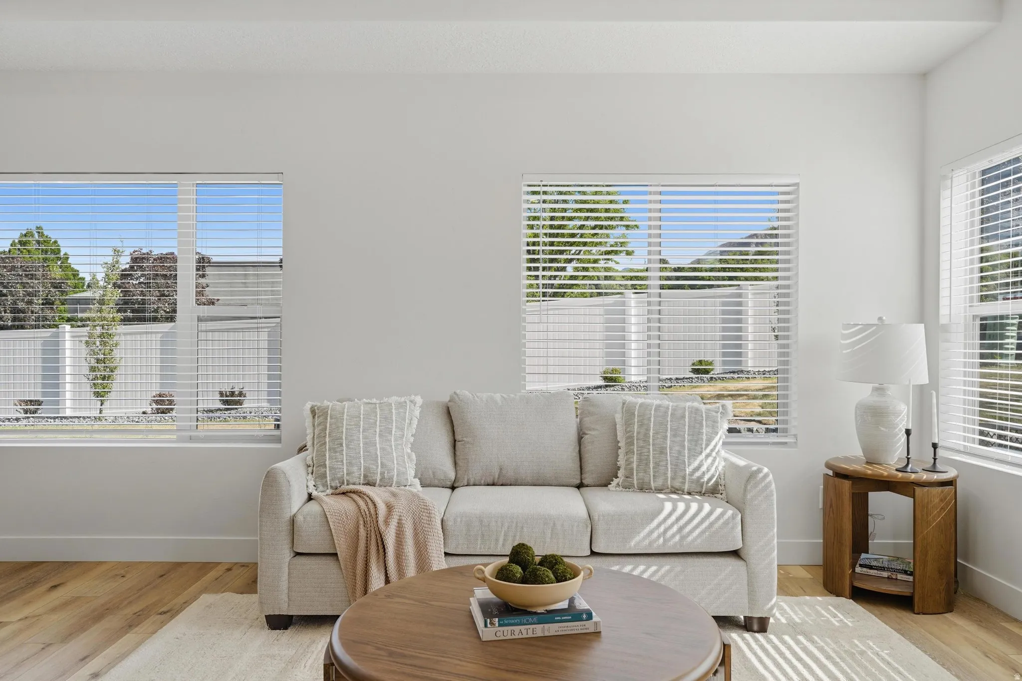 Bright living area featuring white walls, multiple windows with blinds, wood-finish flooring, and a flat ceiling