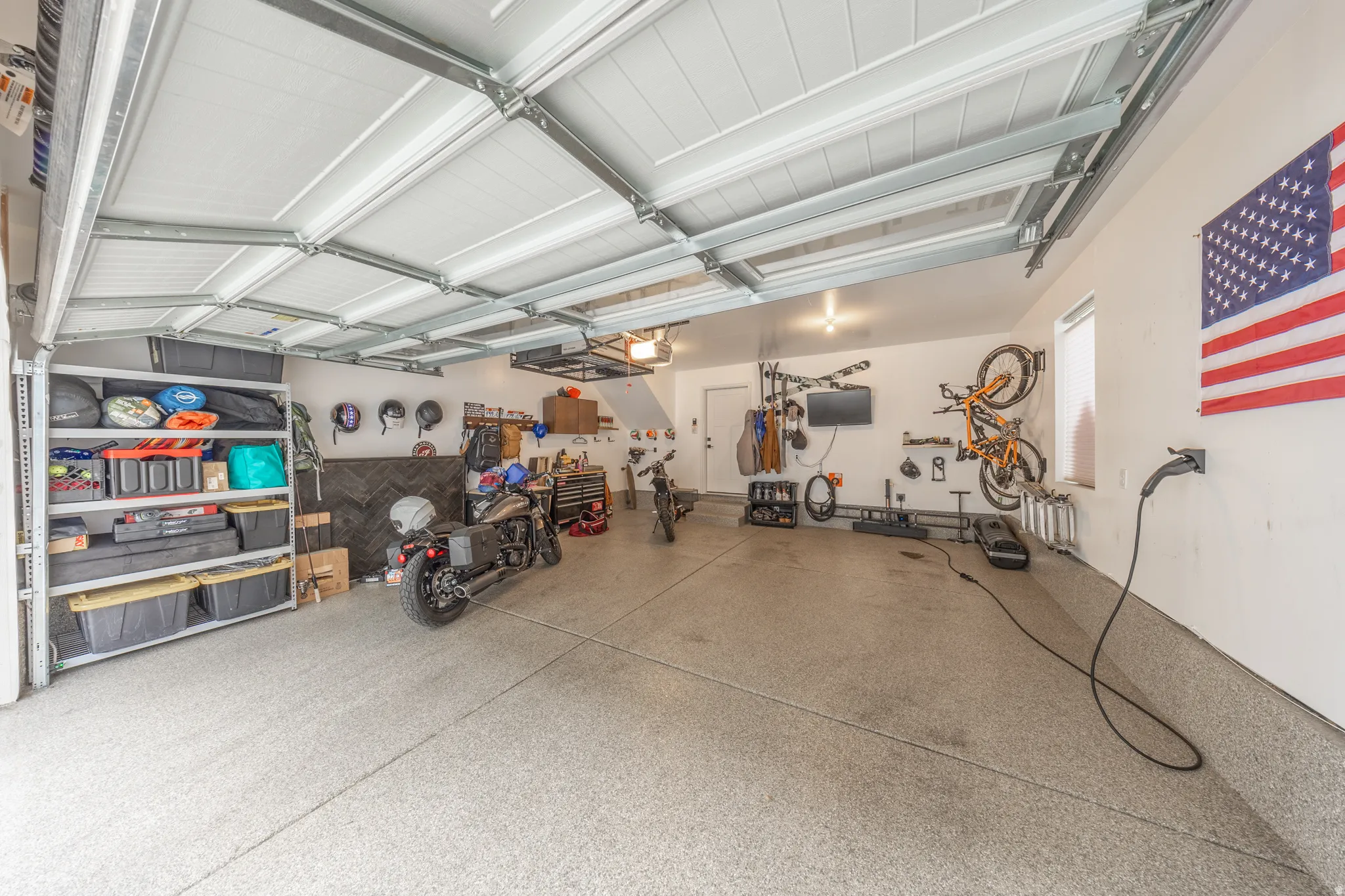 Spacious garage featuring an epoxy-coated concrete floor, ample overhead lighting, an exposed ceiling structure, a dedicated EV charging station, and a built-in storage rack