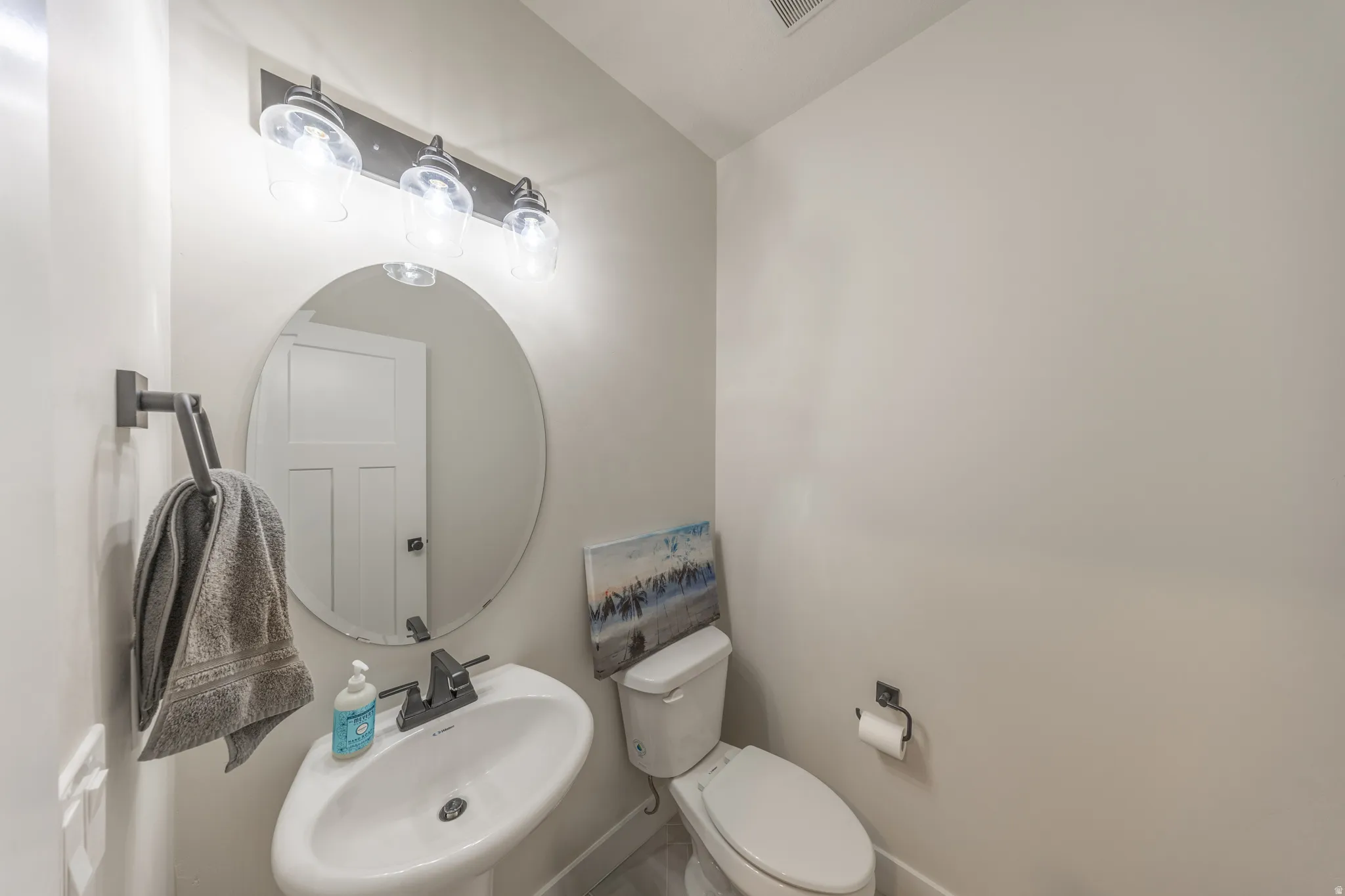 Powder room featuring a pedestal sink, oval mirror, and a three-light vanity fixture with clear glass shades