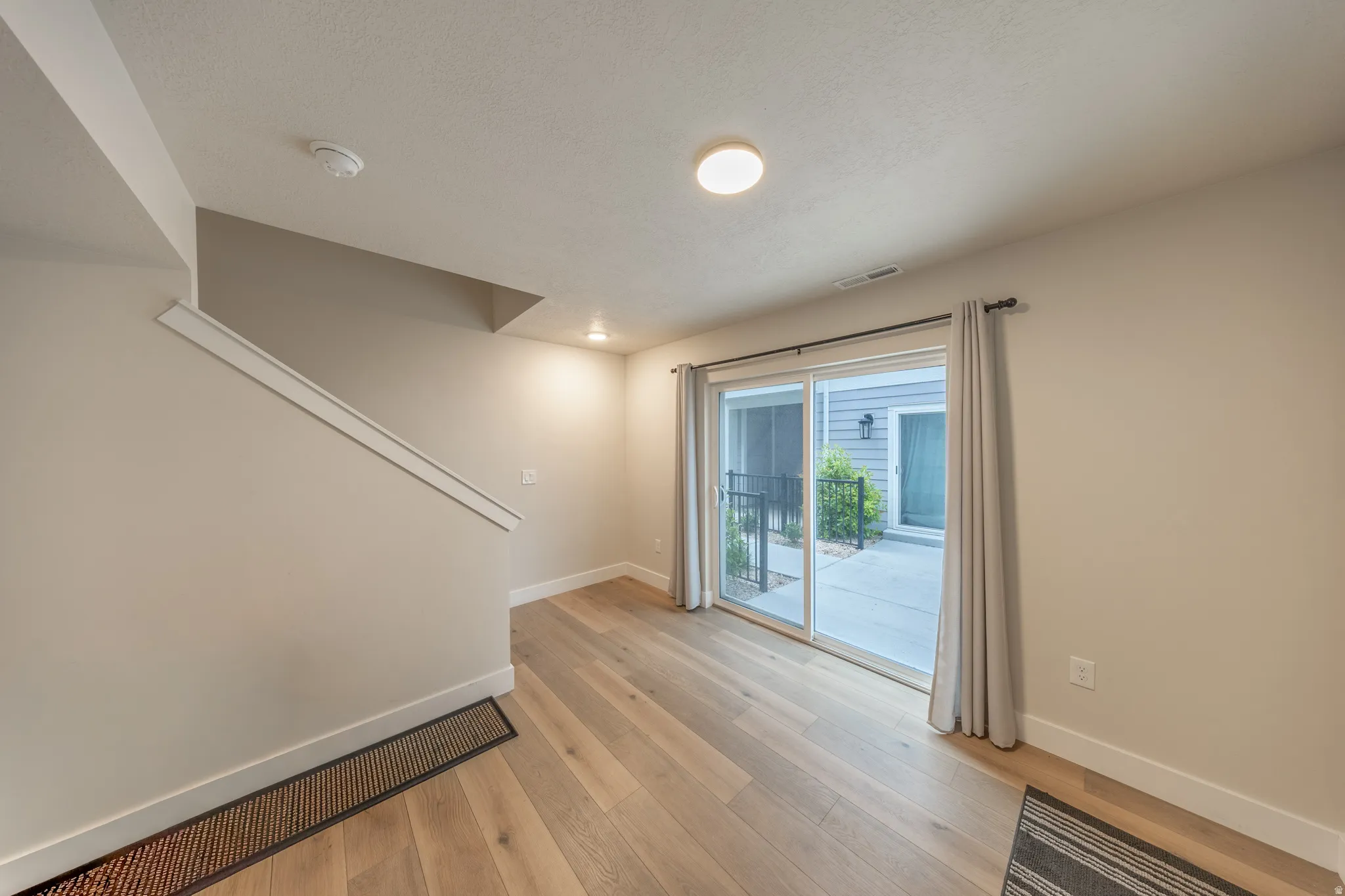 Light wood-finish flooring throughout, featuring a sliding glass door with curtains leading to an outdoor patio with concrete paving and a black metal fence