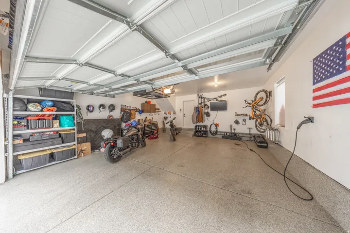 Spacious garage featuring an epoxy-coated concrete floor, ample overhead lighting, an exposed ceiling structure, a dedicated EV charging station, and a built-in storage rack