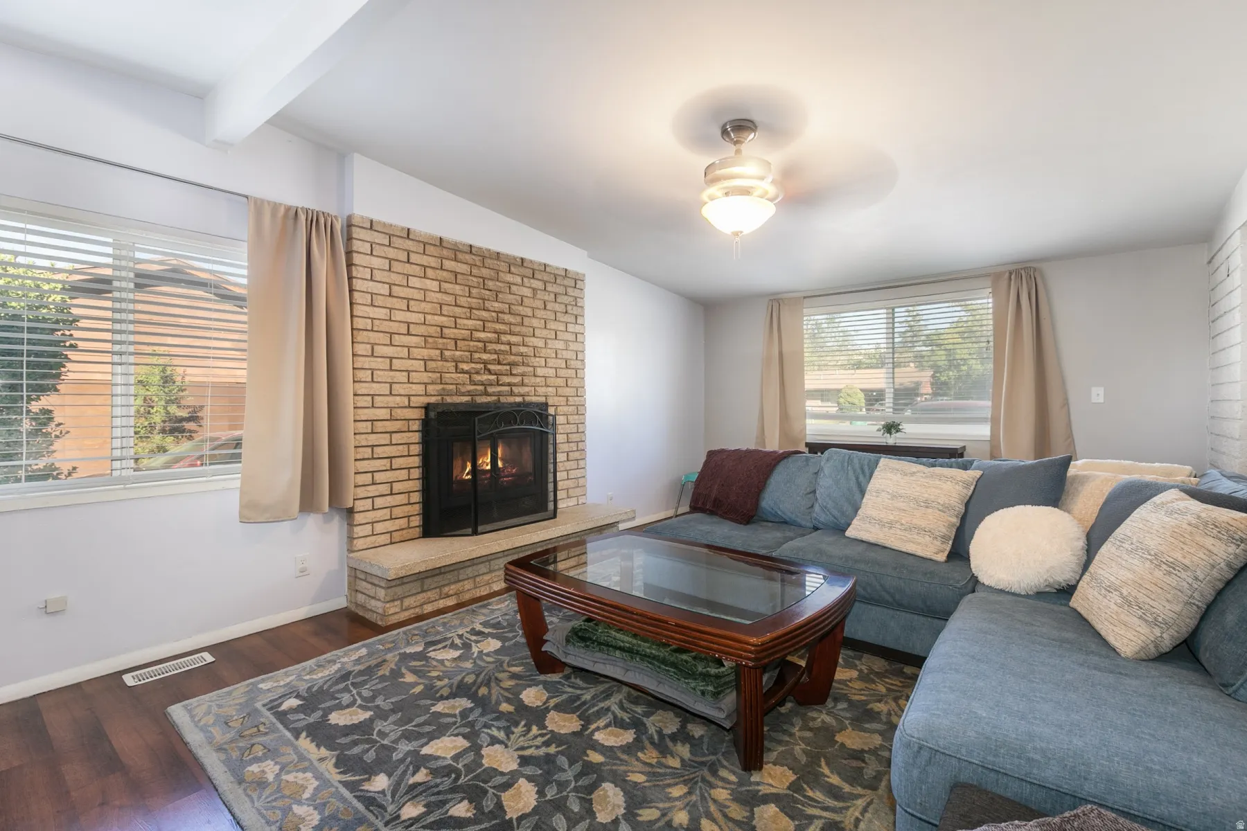Large living or family room right off kitchen with brick-clad fireplace with hearth, wood-finish flooring, ceiling-mounted light fixture, and large windows with blinds