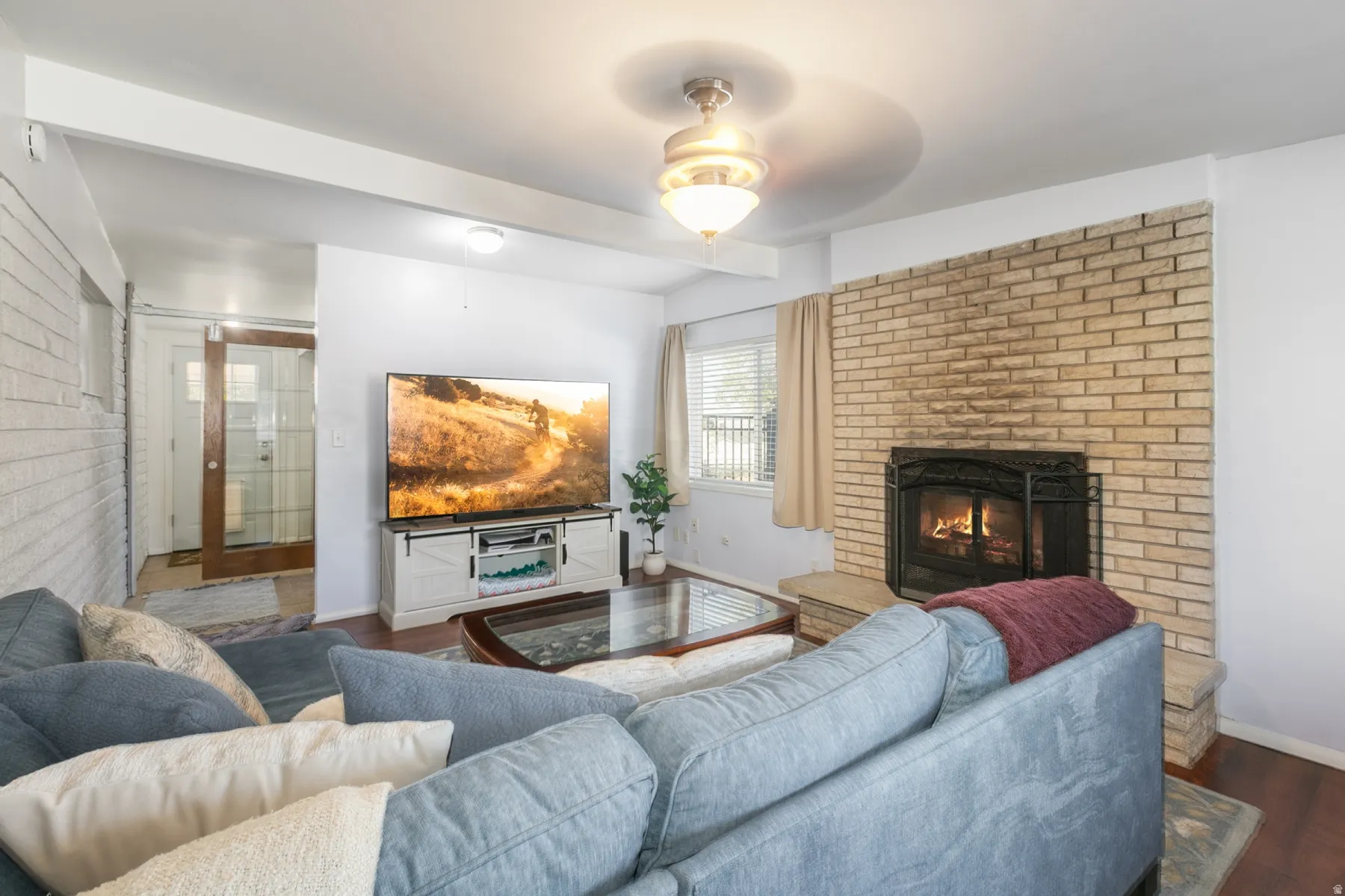 Large living or family room right off kitchen with brick-clad fireplace with hearth, wood-finish flooring, ceiling-mounted light fixture, and large windows with blinds