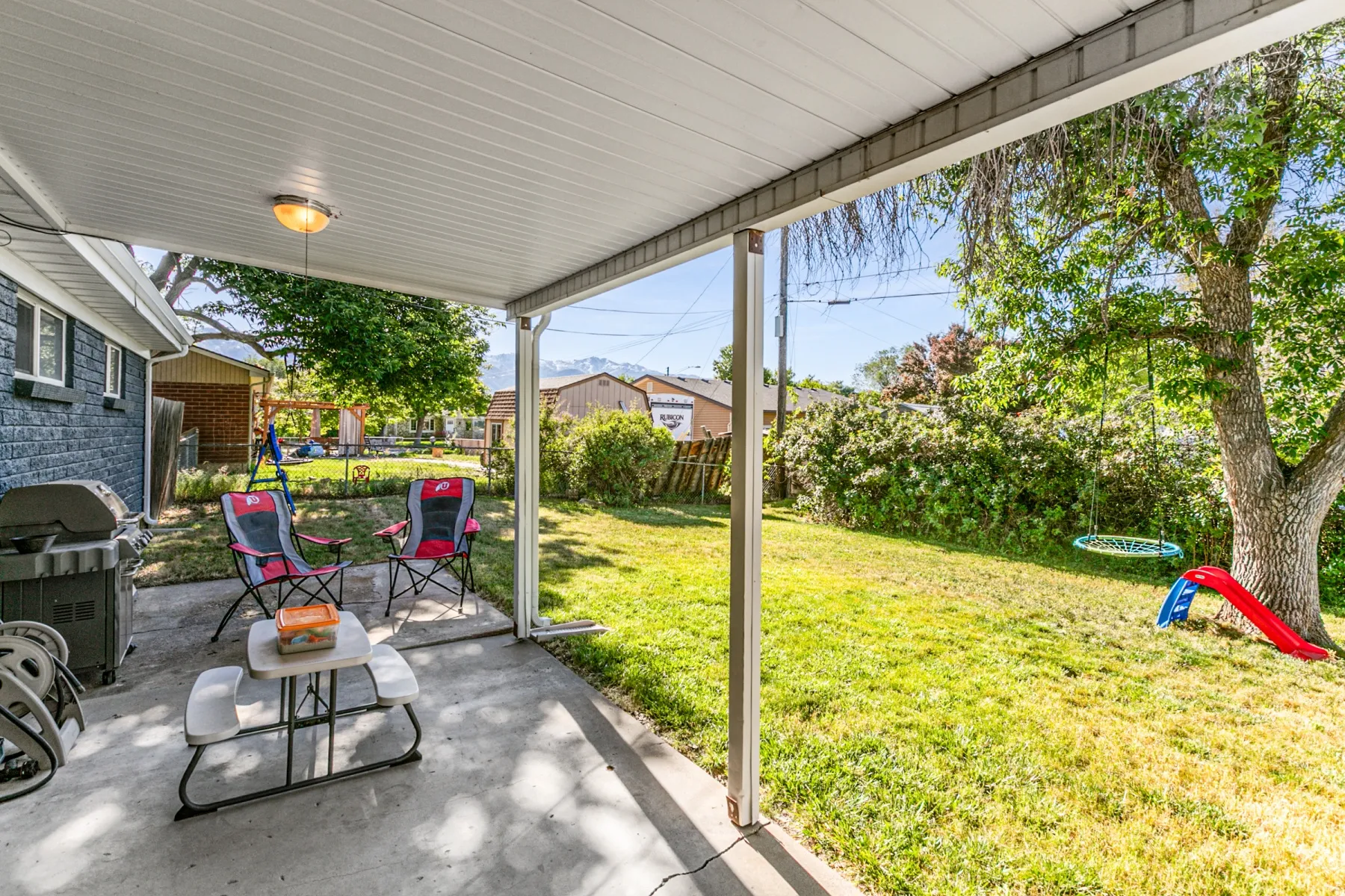 Covered concrete patio with an attached light fixture, featuring a manicured lawn and mature trees in a fully fenced yard.