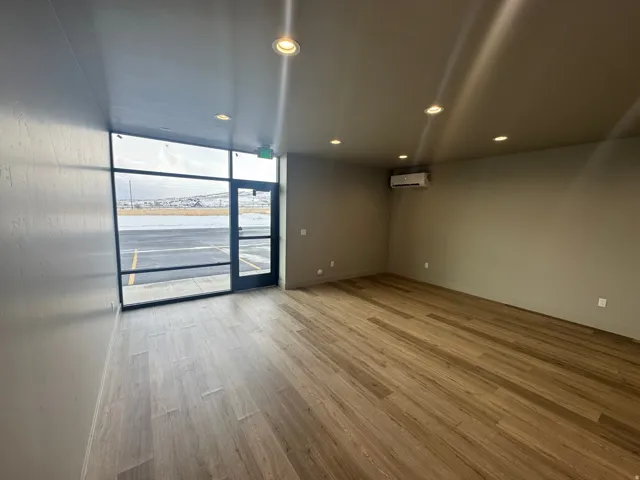 Expansive interior space featuring full-height storefront windows, wood-finish flooring, recessed ceiling lighting, a wall-mounted climate control unit, and a prominent glass entry door