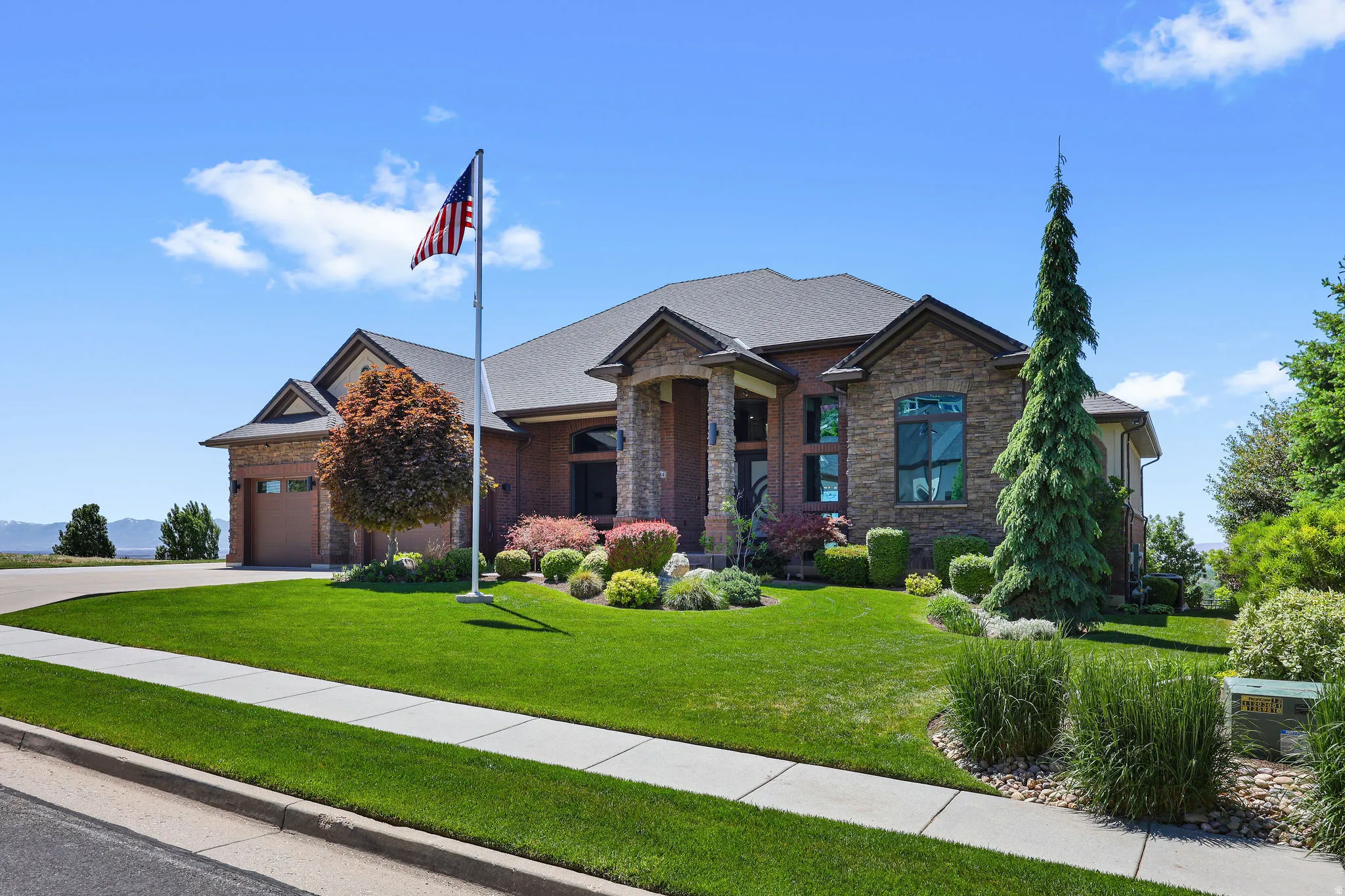Stone and brick-clad home featuring a multi-gabled roofline, an attached garage with a wood-finish door, a manicured lawn, and mature landscaping