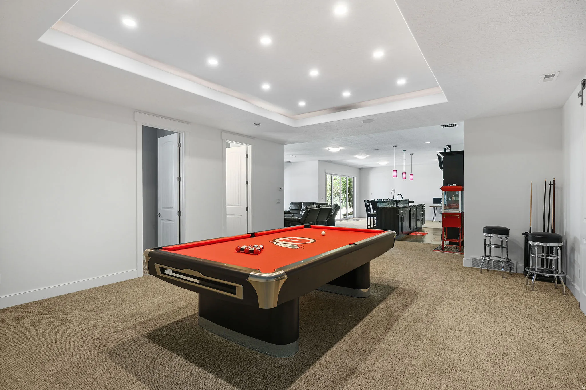 Expansive recreation room featuring a neutral carpet flooring, recessed tray ceiling with ample lighting, and an open layout to an additional living area