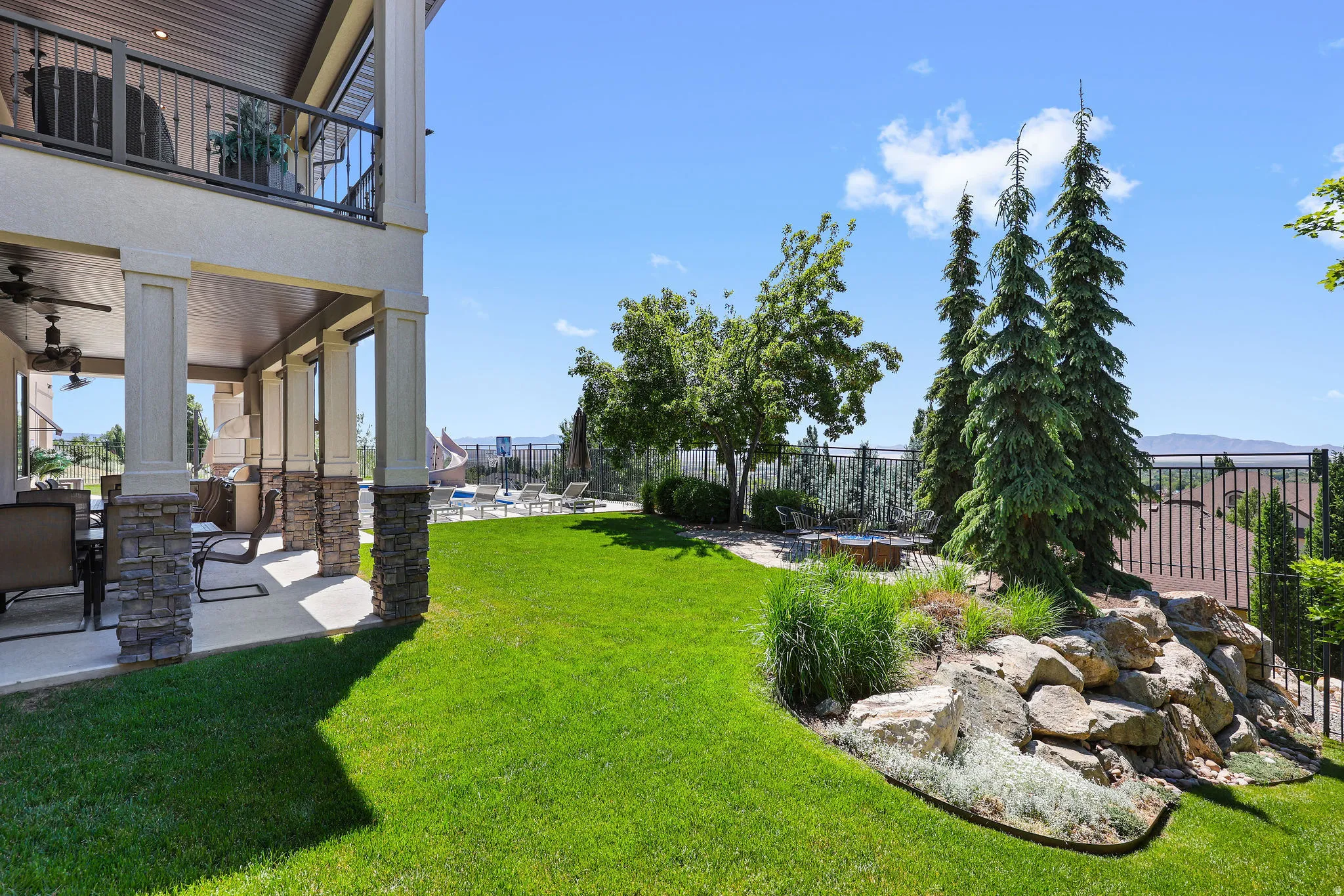 Expansive rear yard featuring a manicured lawn, tiered landscaping with mature evergreens and natural rock, and a fenced perimeter