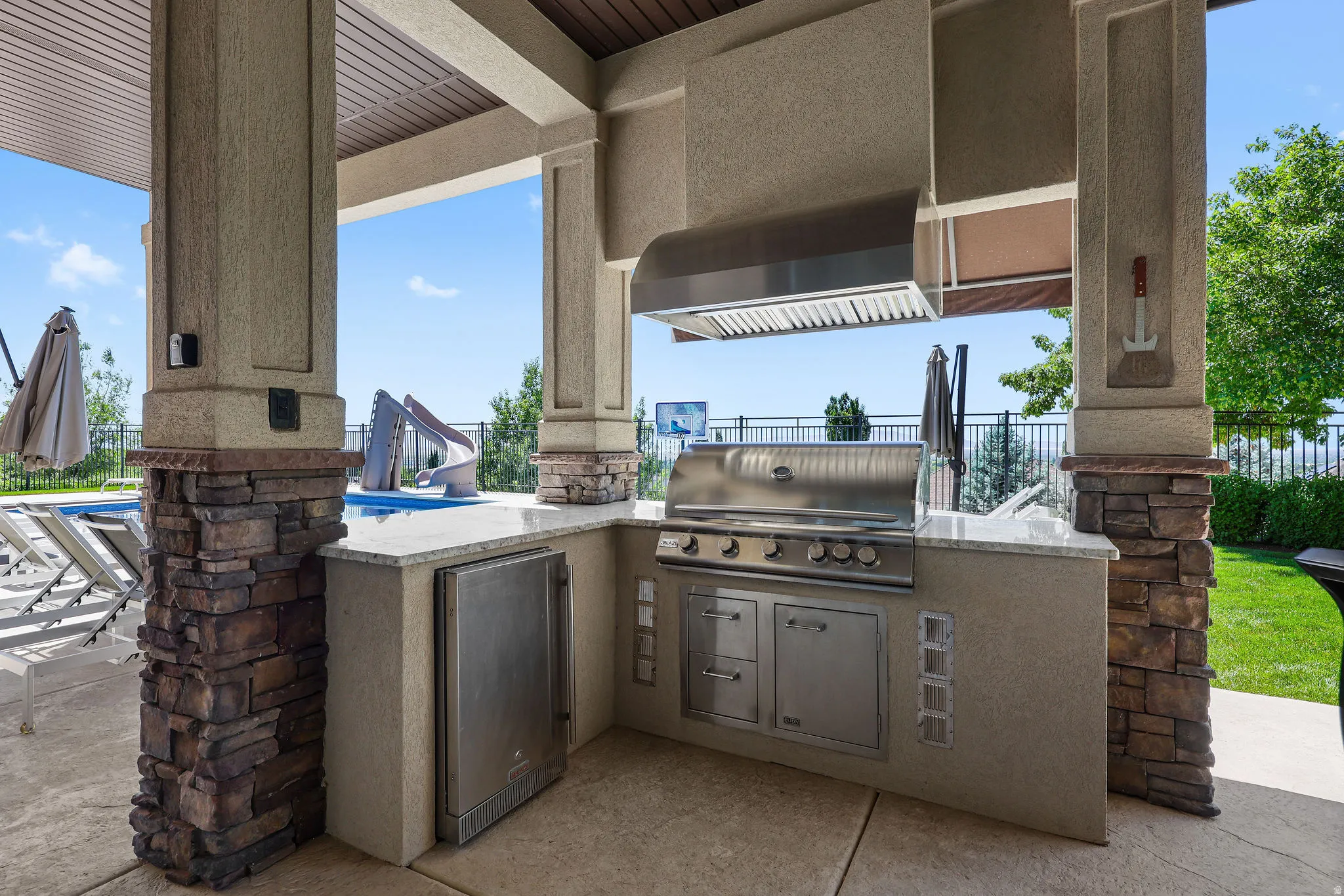 Outdoor kitchen featuring a built-in stainless steel grill, stainless steel exhaust hood, built-in stainless steel refrigerator, light-toned countertop, and stone-accented columns