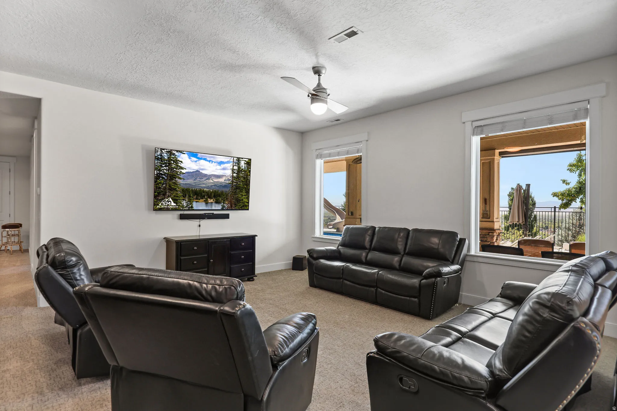 Carpeted entertainment room featuring white walls, a ceiling fan with integrated lighting, and two large windows