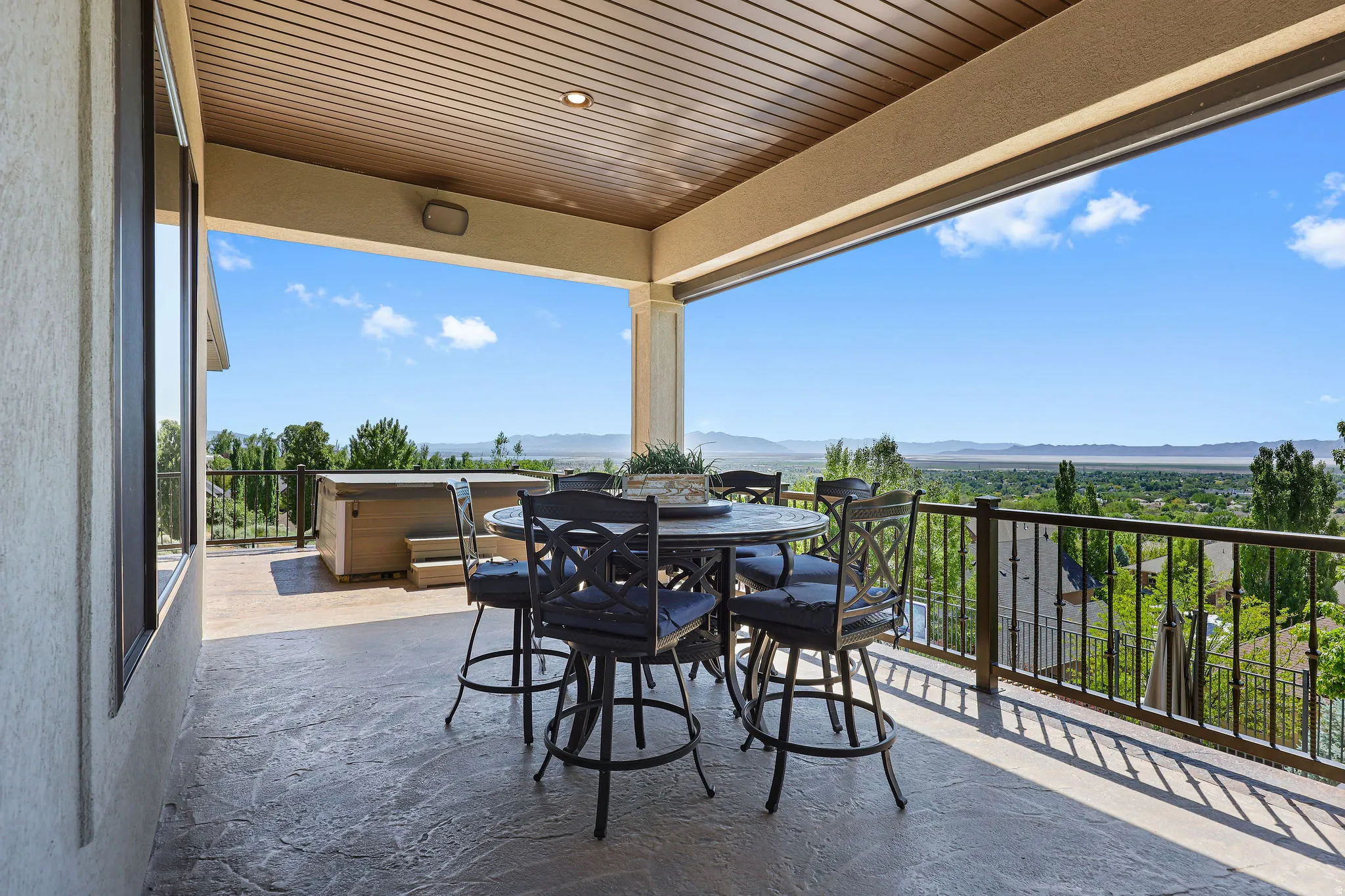 Covered patio featuring stamped concrete flooring, a wood-finish ceiling with recessed lighting, an exterior hot tub, and a black wrought iron railing overlooking distant mountains