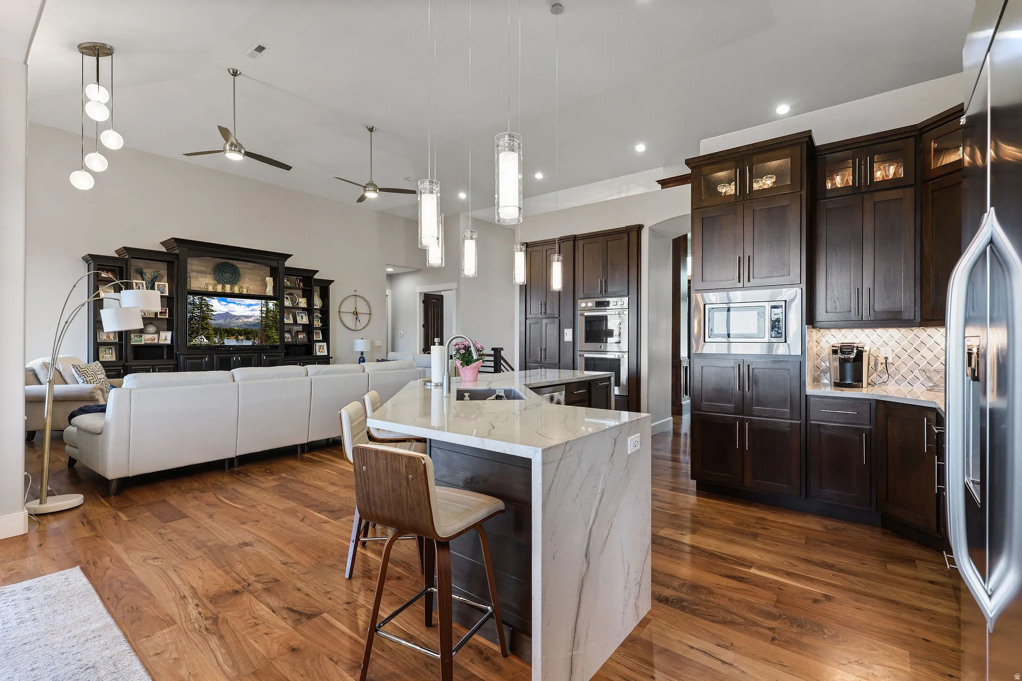 Open-concept living space featuring wood-finish flooring throughout, a kitchen island with a waterfall countertop, dark wood cabinetry with integrated stainless steel appliances, and various ceiling light fixtures