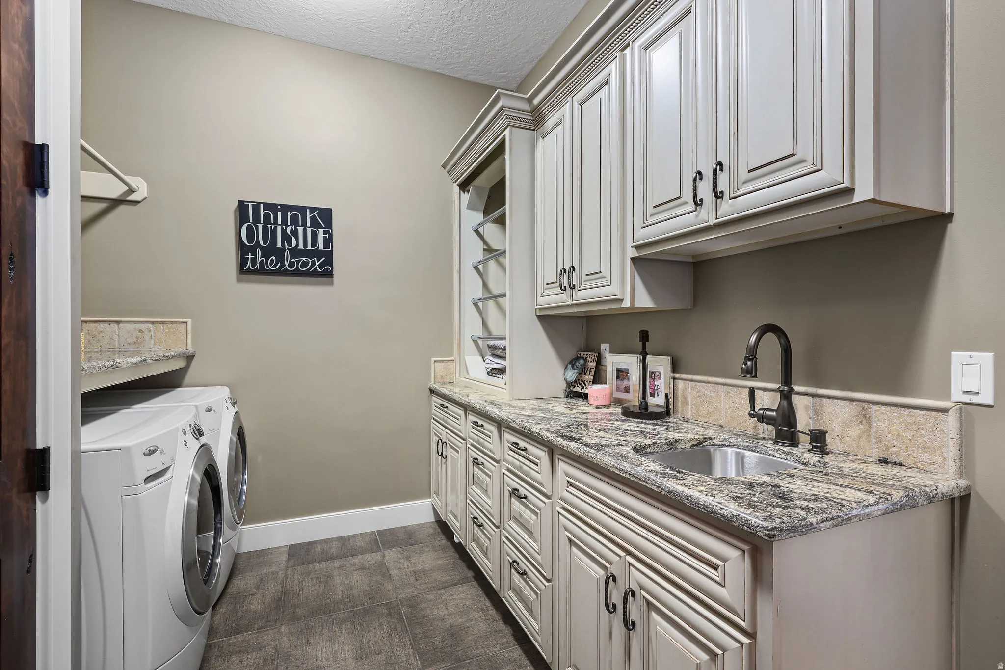 Laundry room featuring extensive custom cabinetry with distressed white finish, a stainless steel sink with a dark bronze faucet, and a stone countertop with a matching backsplash
