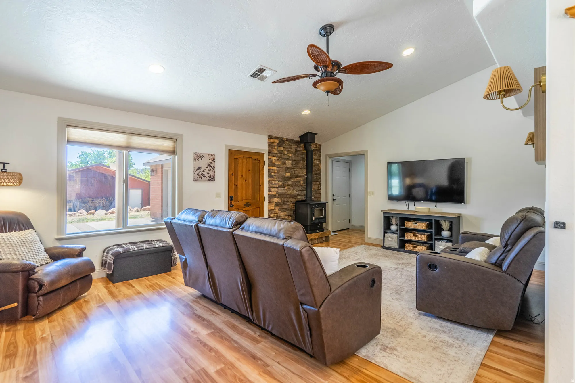 Bright living space featuring wood-finish flooring, a stone accent wall with a wood-burning stove, and a vaulted ceiling with a ceiling fan