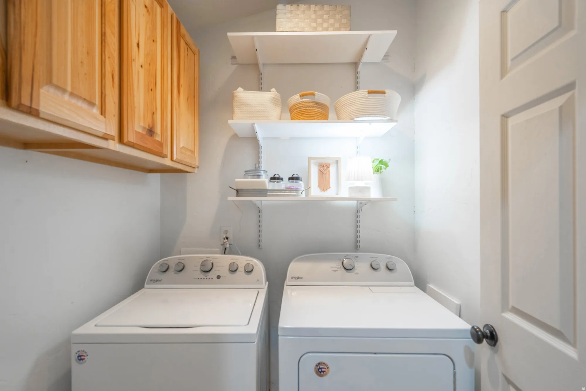 Dedicated utility room featuring wood-finish cabinetry, white wall shelving, light-toned wall paint, and a white paneled door with a matte black doorknob
