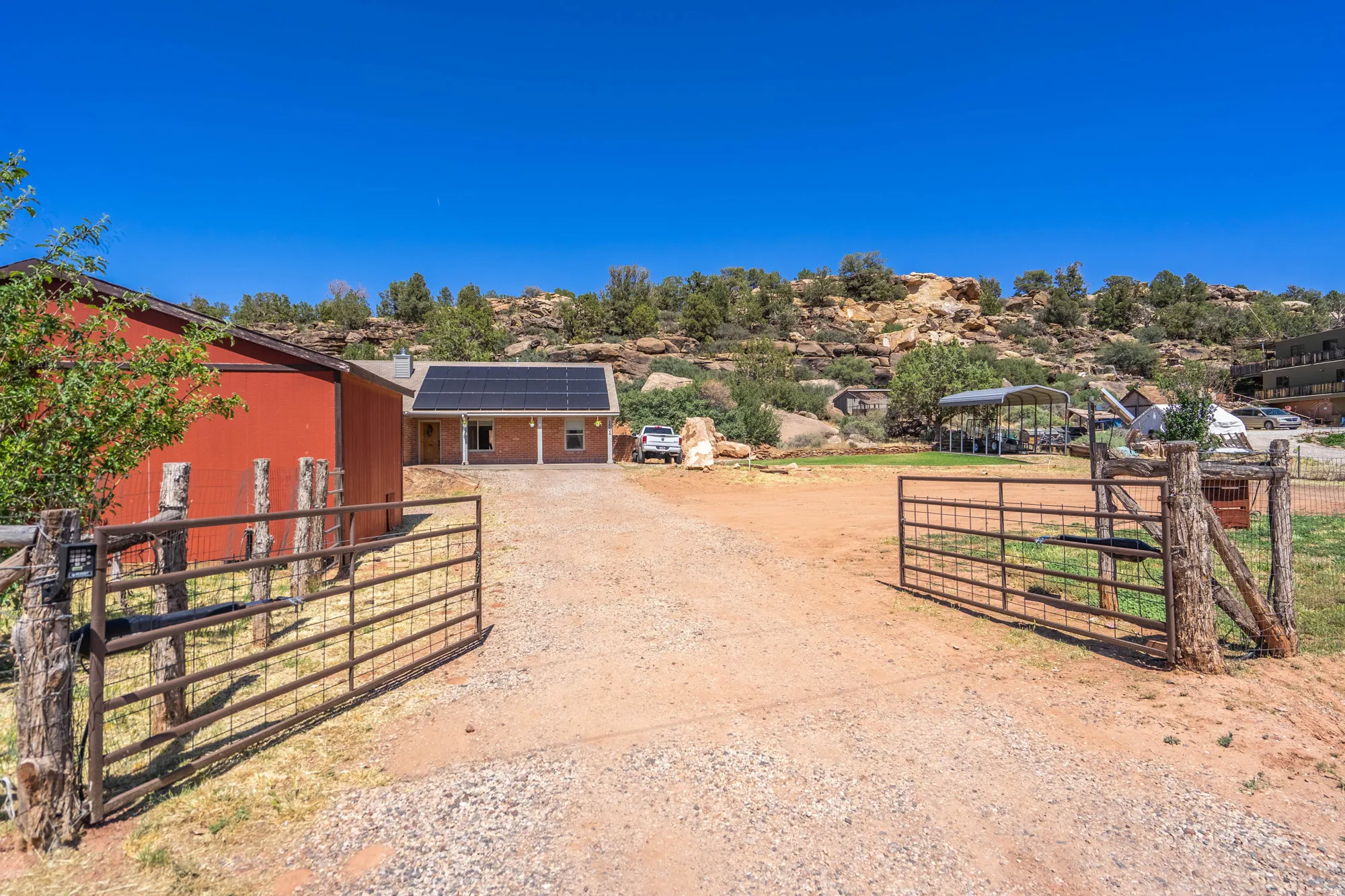 Gated gravel driveway with a rustic timber fence, red barn, and a residence featuring solar panels and an extended covered porch