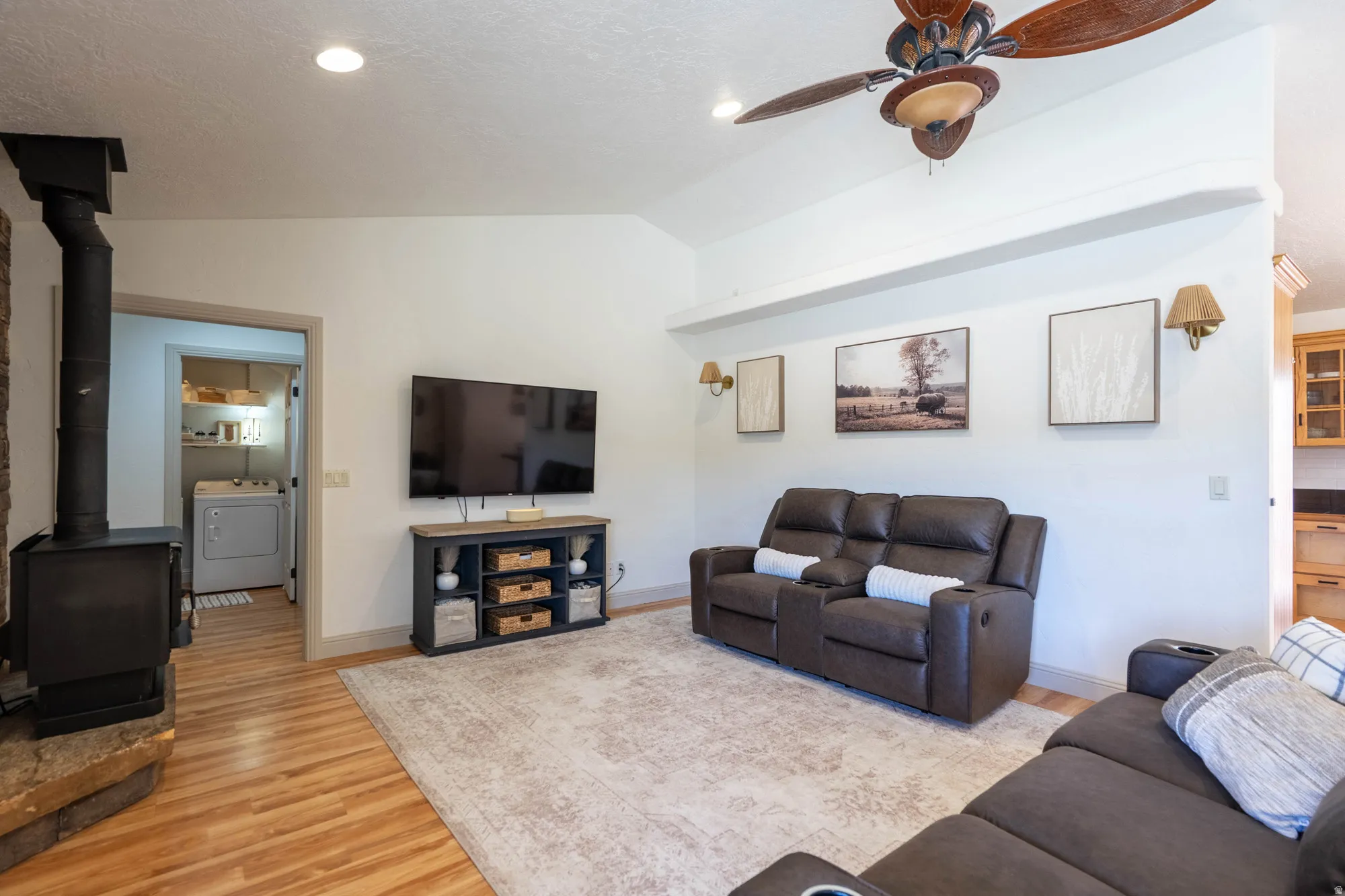 Vaulted ceilings with a ceiling fan, wood-finish flooring, recessed lighting, and a wood-burning stove with a stone surround
