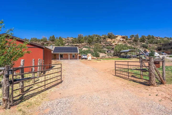 Gated gravel driveway with a rustic timber fence, red barn, and a residence featuring solar panels and an extended covered porch