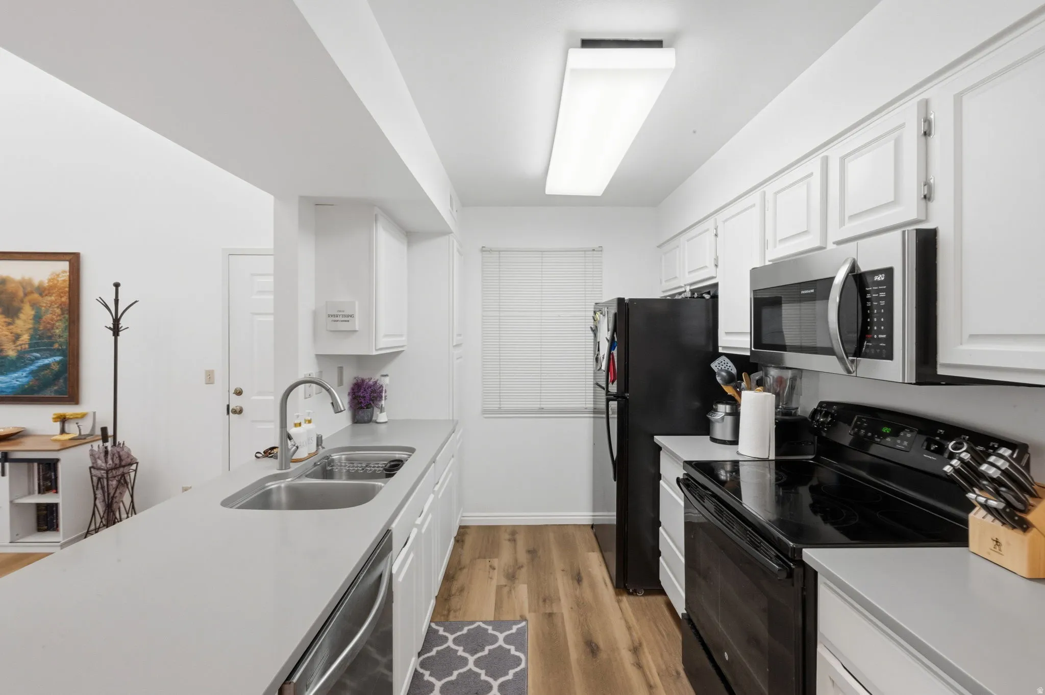 Galley kitchen featuring light wood-finish flooring, white cabinetry, a double basin stainless steel sink, light grey countertops, and a full suite of black and stainless steel appliances