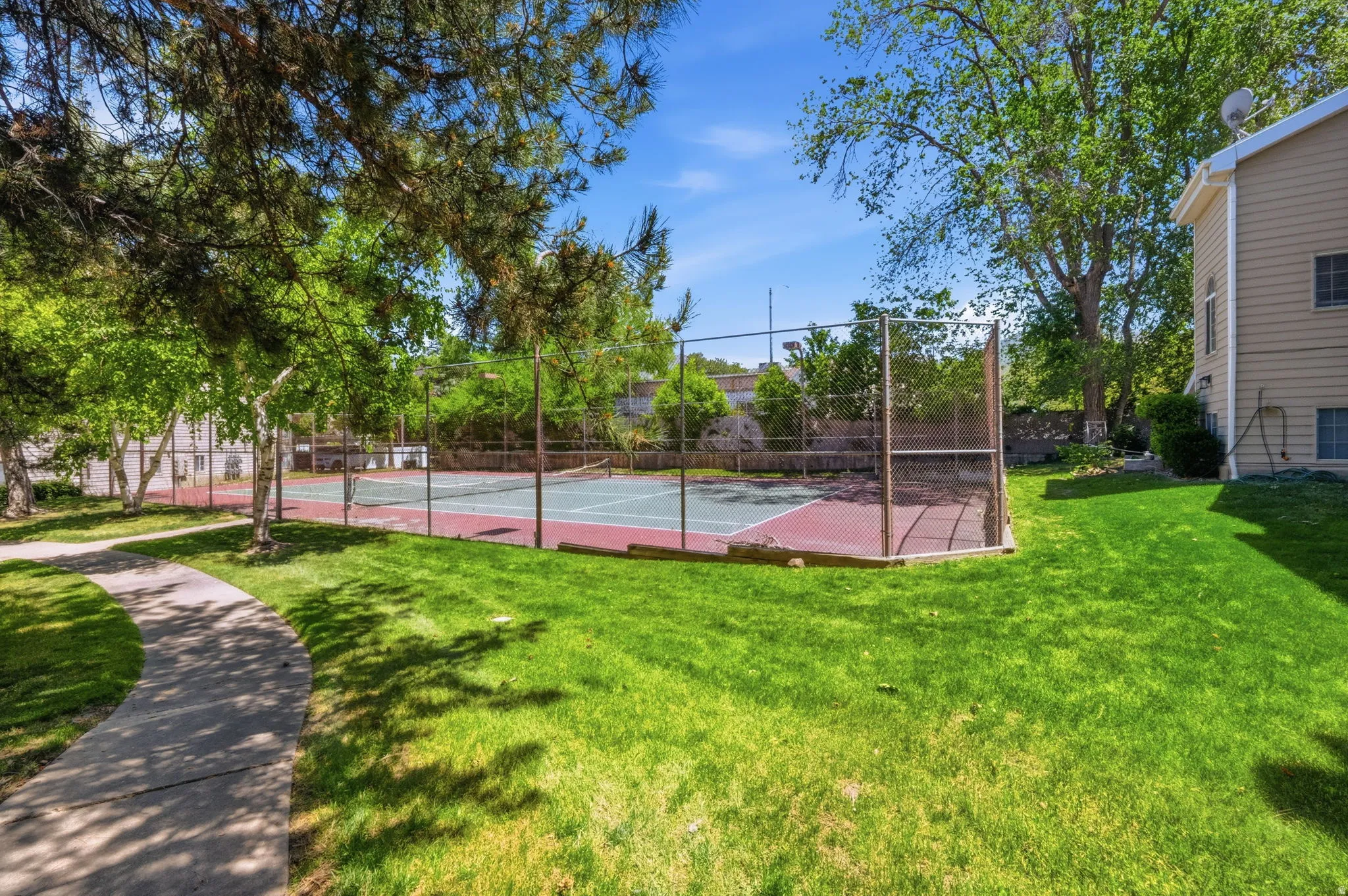 Outdoor tennis court with chain-link fencing, surrounded by mature trees and a manicured lawn