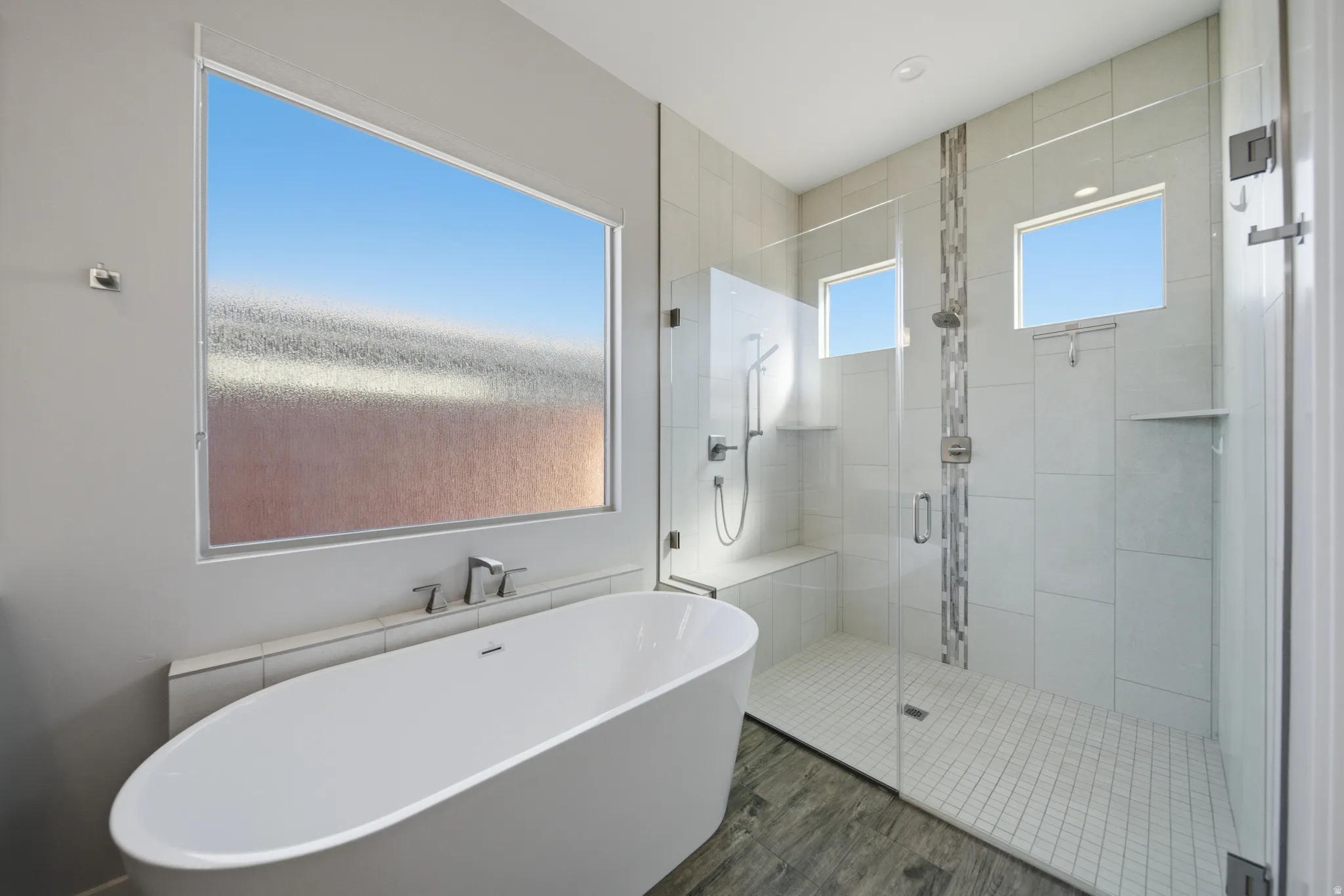 Bathroom featuring a freestanding oval tub with deck-mounted fixtures, a spacious glass-enclosed shower, light-toned tile walls, wood-finish flooring, and recessed lighting