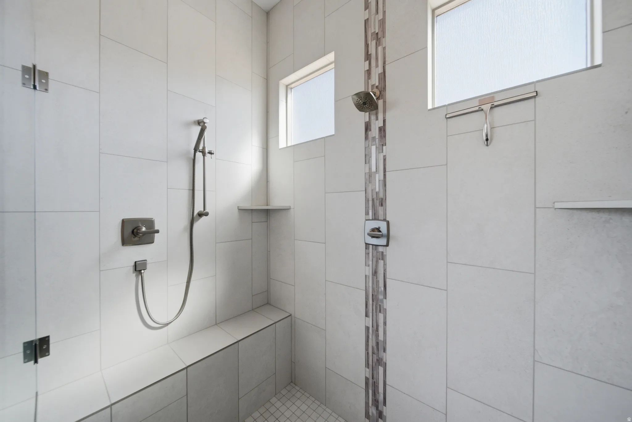 Spacious tiled shower featuring a built-in bench, dual recessed shelves, a handheld showerhead, a rainfall showerhead, and translucent horizontal windows