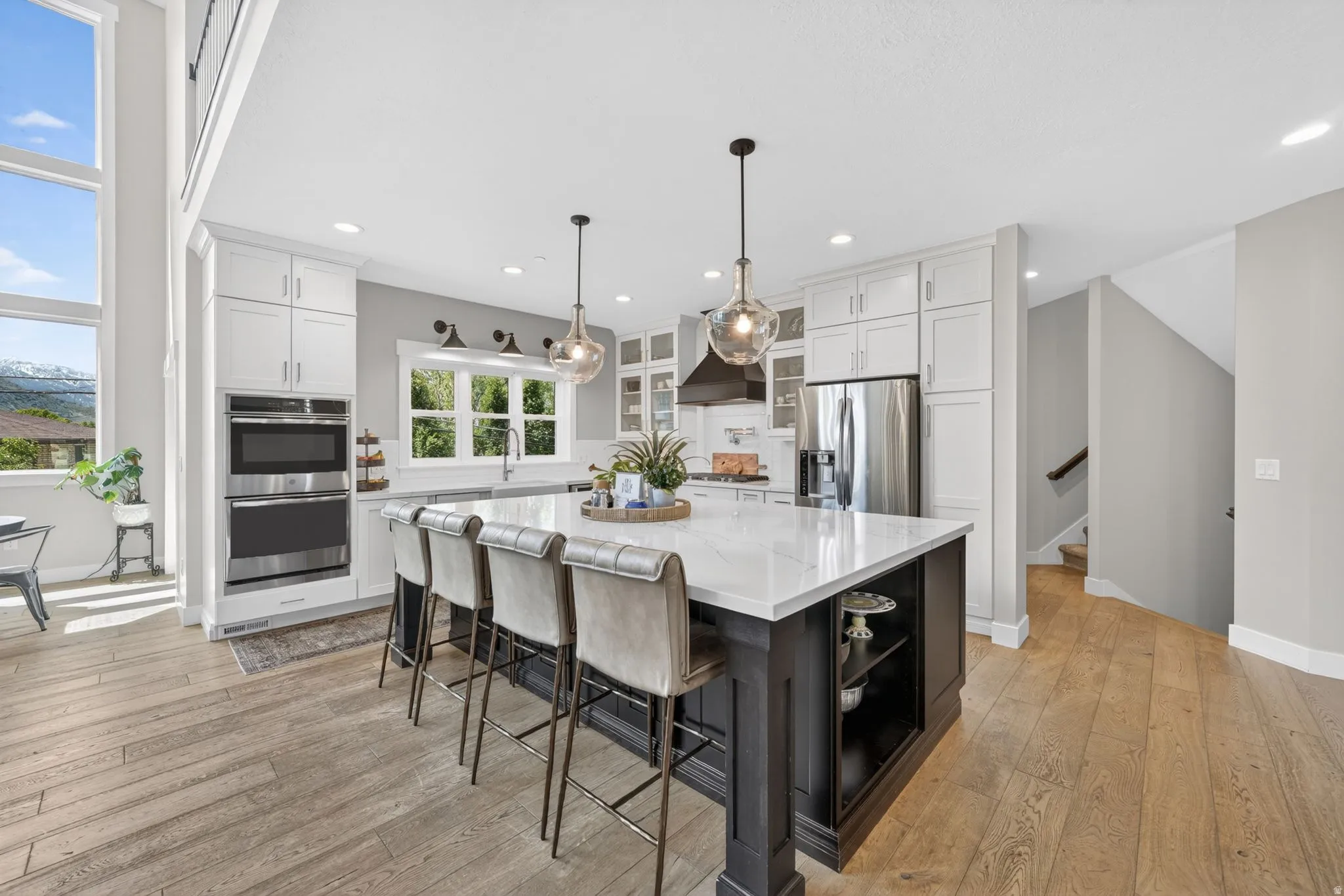 Open-concept kitchen featuring a dark-toned island with a white countertop, light wood-finish flooring, white cabinetry, stainless steel appliances, and large windows providing abundant illumination