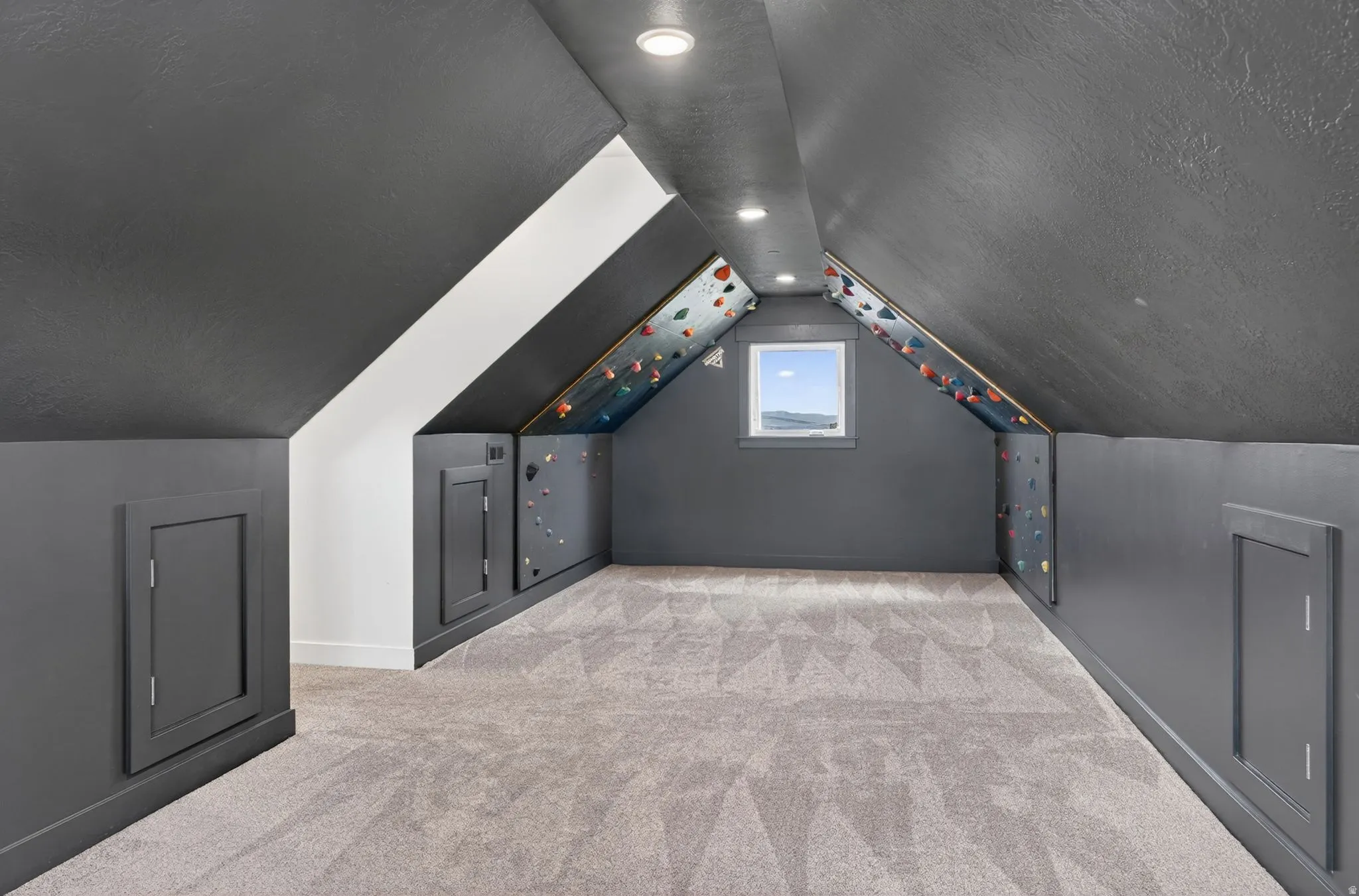 Vaulted ceiling room featuring recessed lighting, a window, built-in storage access panels, and wall-mounted climbing holds
