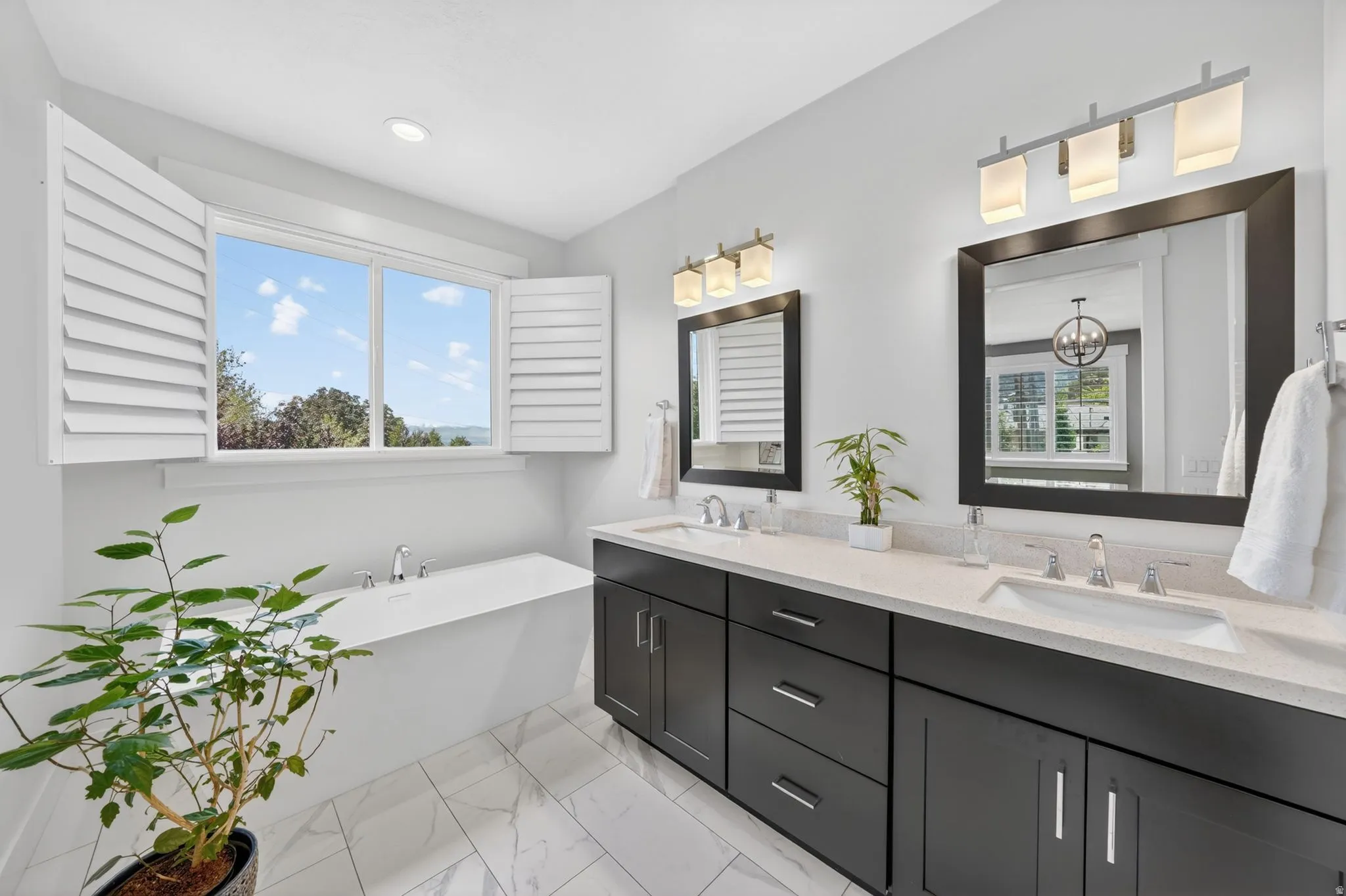 Contemporary bathroom featuring a freestanding soaking tub, a dual vanity with undermount sinks, stone countertops, dark cabinetry, and white plantation shutters
