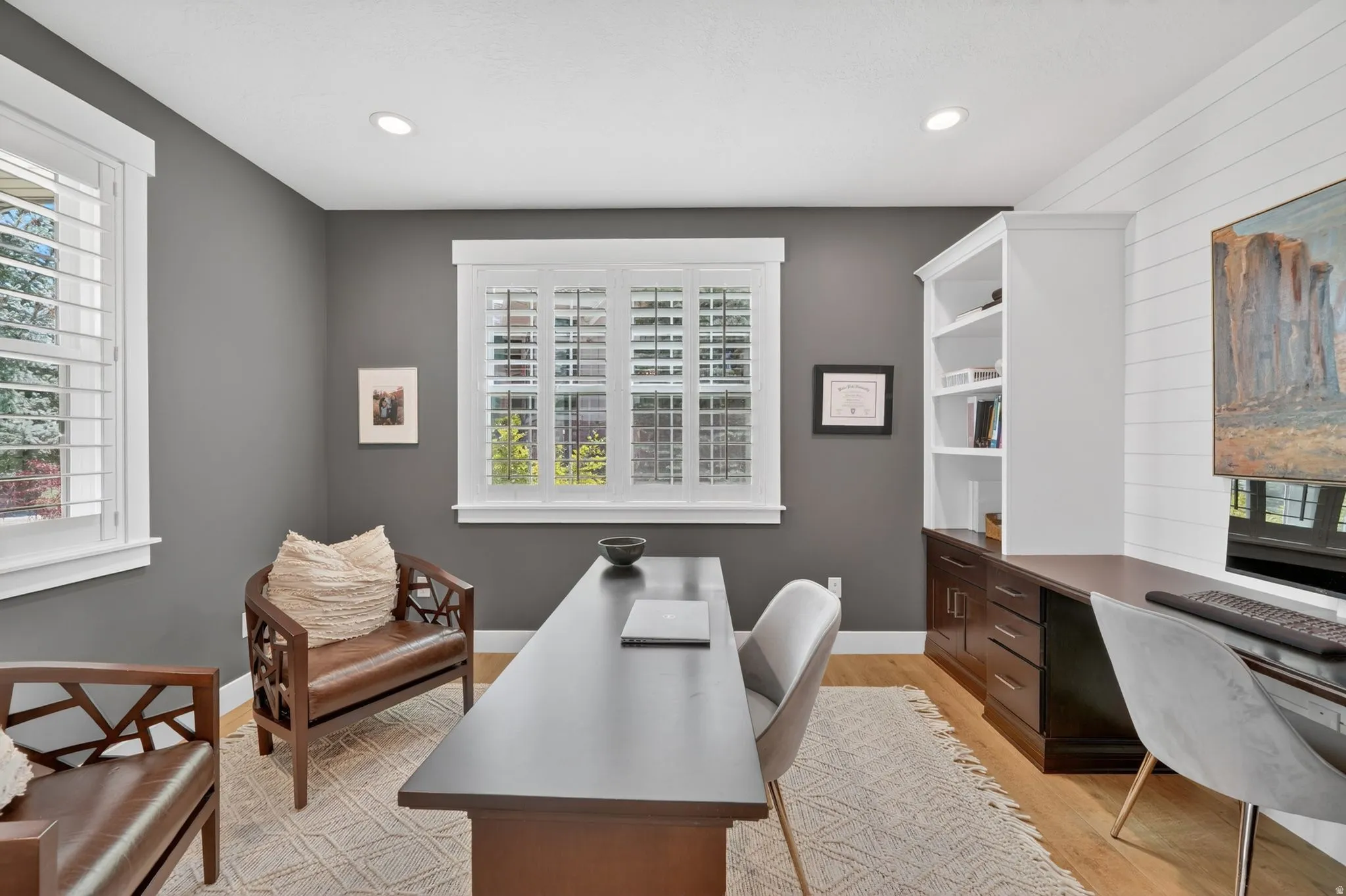Dedicated office space featuring two windows with white plantation shutters, built-in shelving, integrated desk, and wood-finish flooring