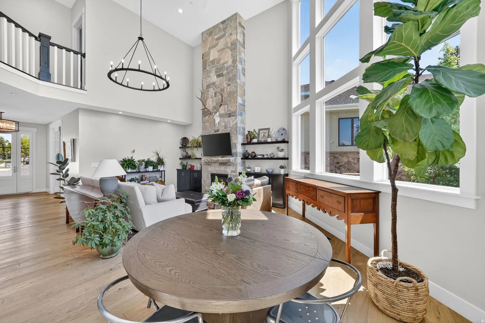 Two-story living area featuring a stone fireplace, high ceilings, large windows, light wood-finish flooring, and a second-story balcony with dark wood and white baluster railing