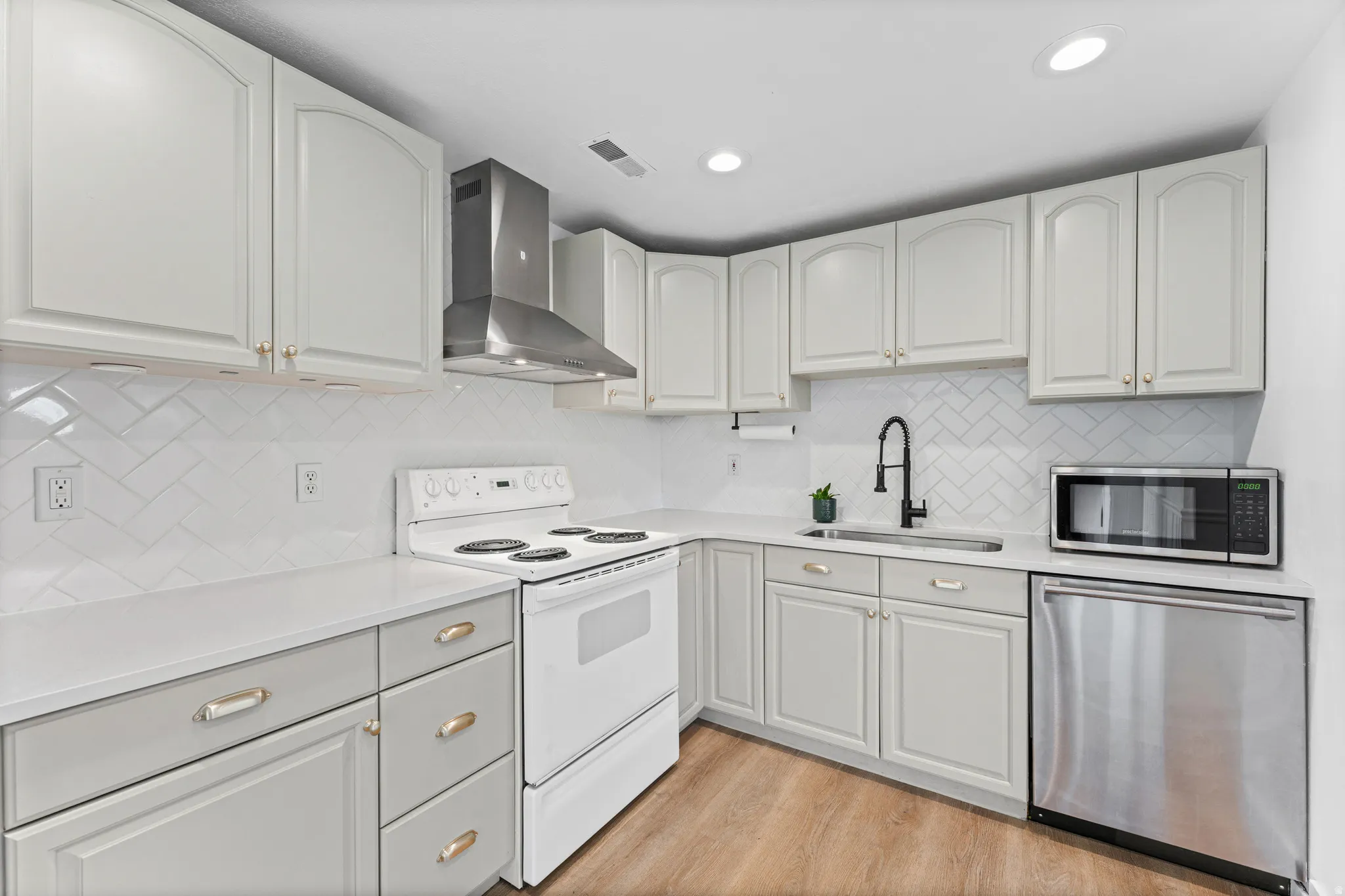 Well-appointed kitchen featuring light-toned cabinetry, a herringbone tile backsplash, white countertops, a stainless steel range hood, and wood-finish flooring
