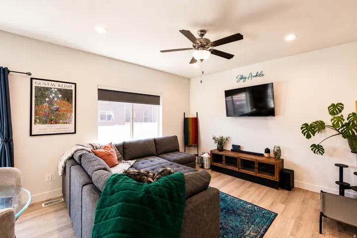 Spacious living area featuring light wood-finish flooring, a contemporary ceiling fan with integrated lighting, recessed lighting, and a large window with a roller shade