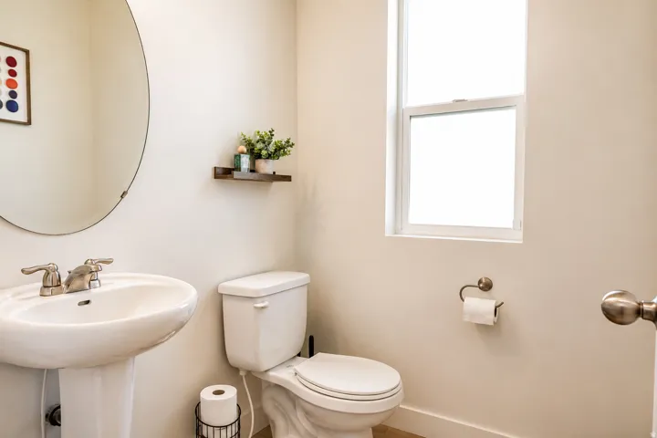 Pedestal sink with chrome-finish faucet, circular wall mirror, and toilet with a window above